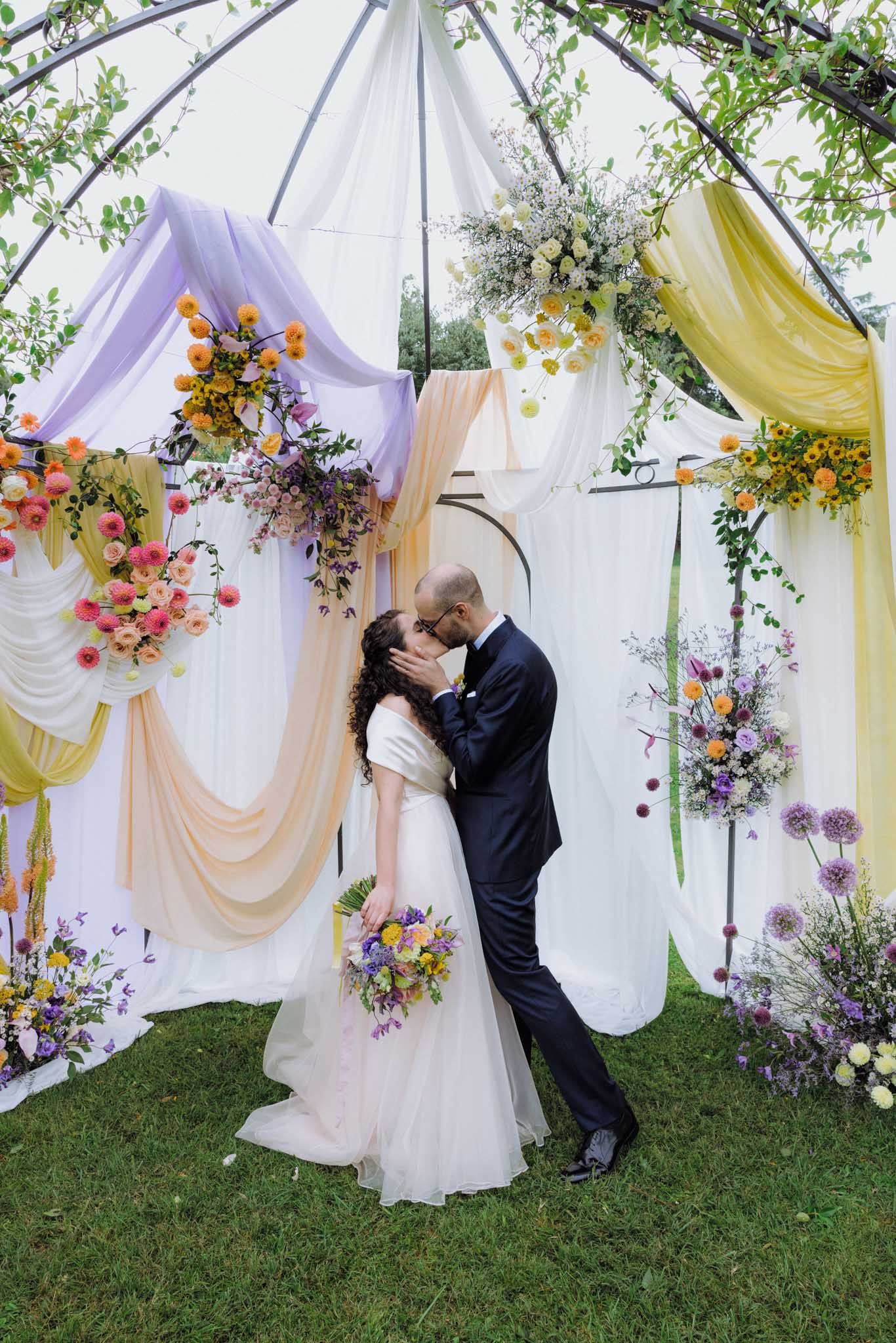 Romantic outdoor wedding ceremony setup with draped fabric in purple and yellow with floral arrangements under a canopy.