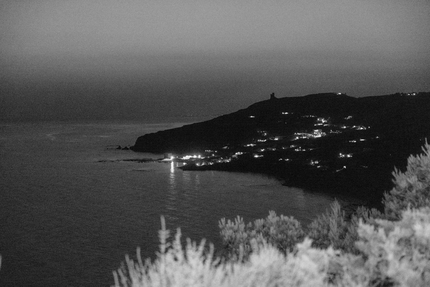 Black and white coastal landscape with twinkling lights from a seaside village and lighthouse on a dark hillside.