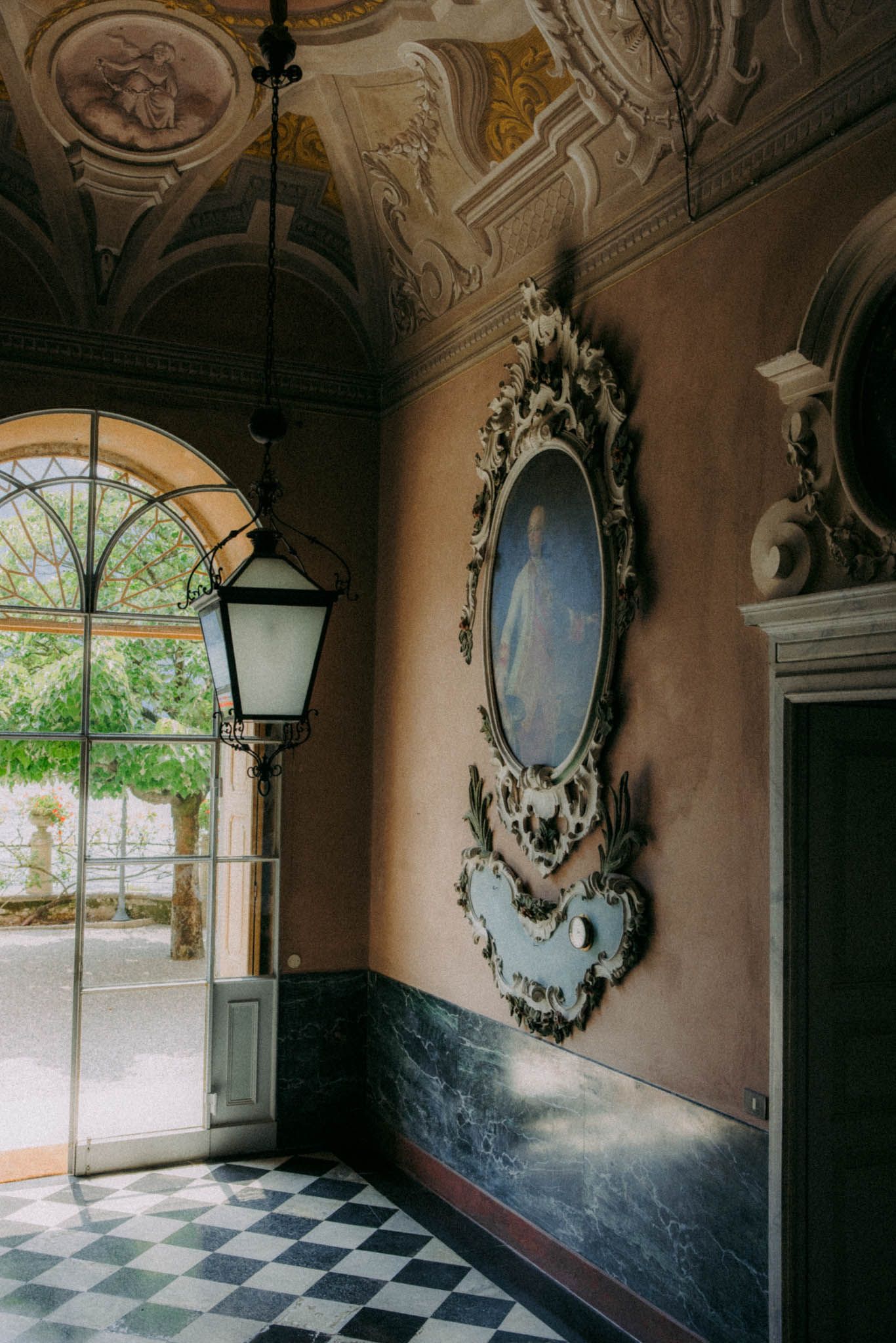 Ornate hallway with checkerboard floor and antique mirrors features elegant baroque-style decor and natural light from windows.