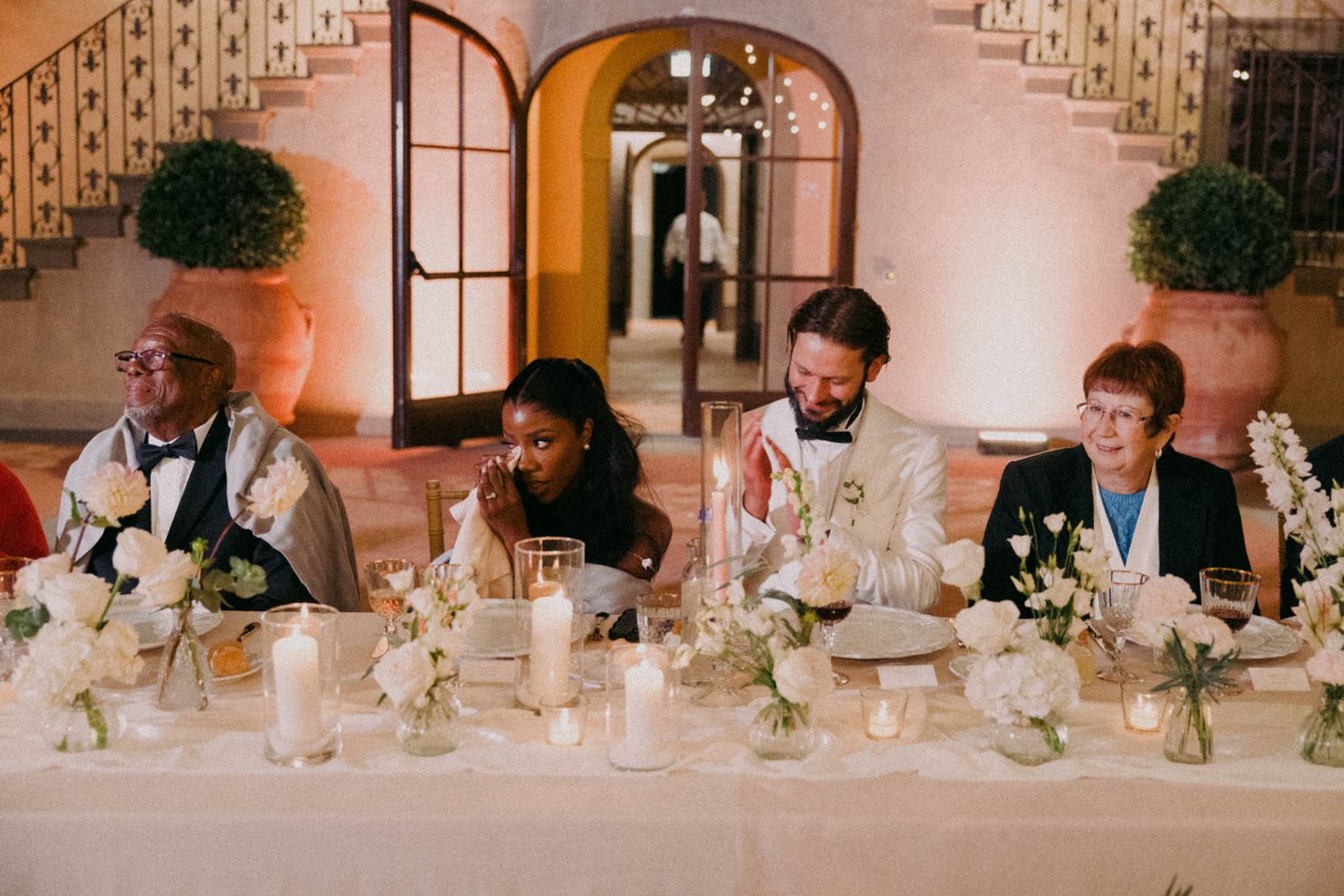 Wedding guests seated at a long table decorated with white linens and floral centerpieces during a reception celebration.