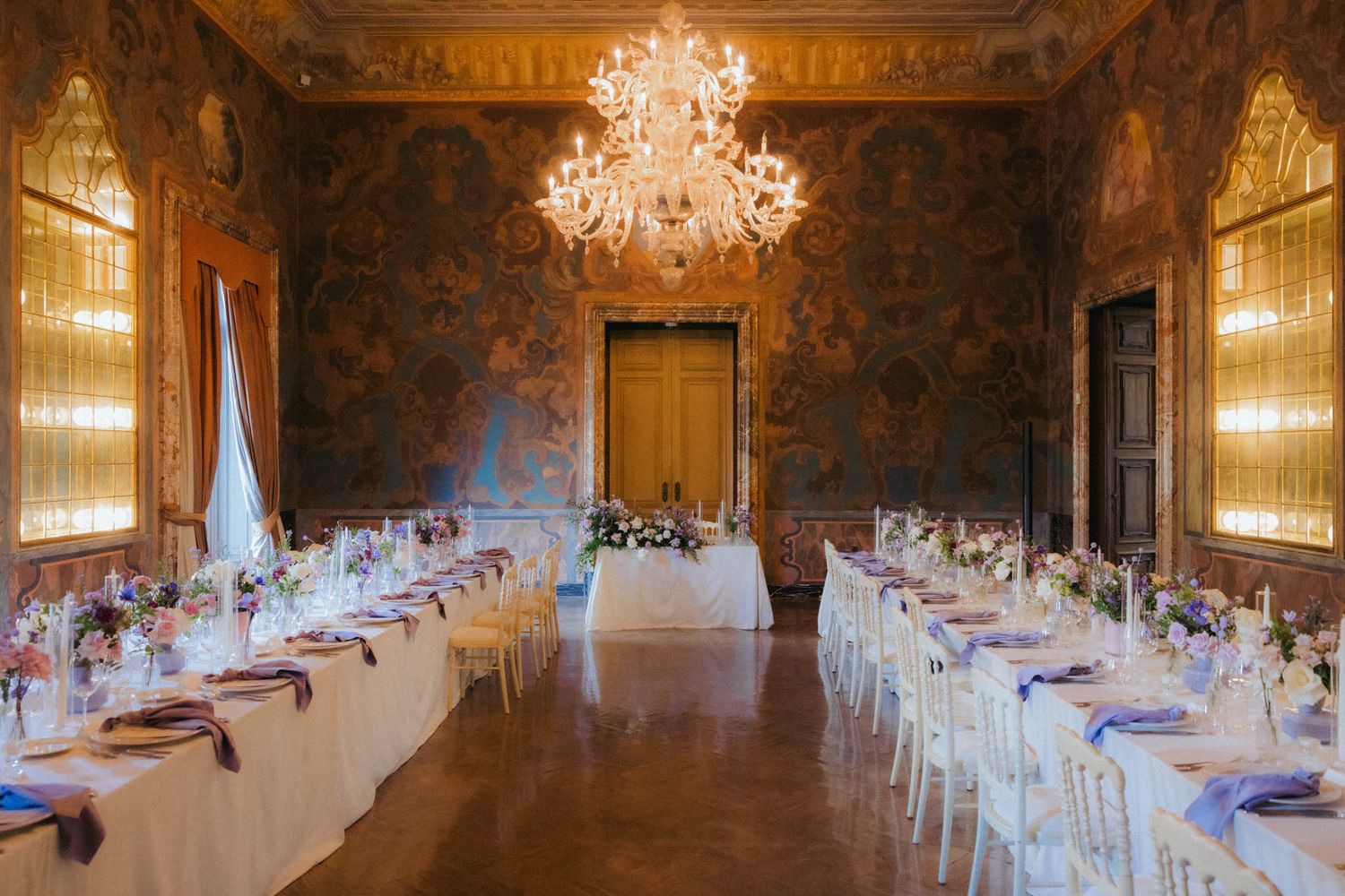 Elegant banquet hall with crystal chandelier and long white tables set for formal dining in ornate room with gold walls.