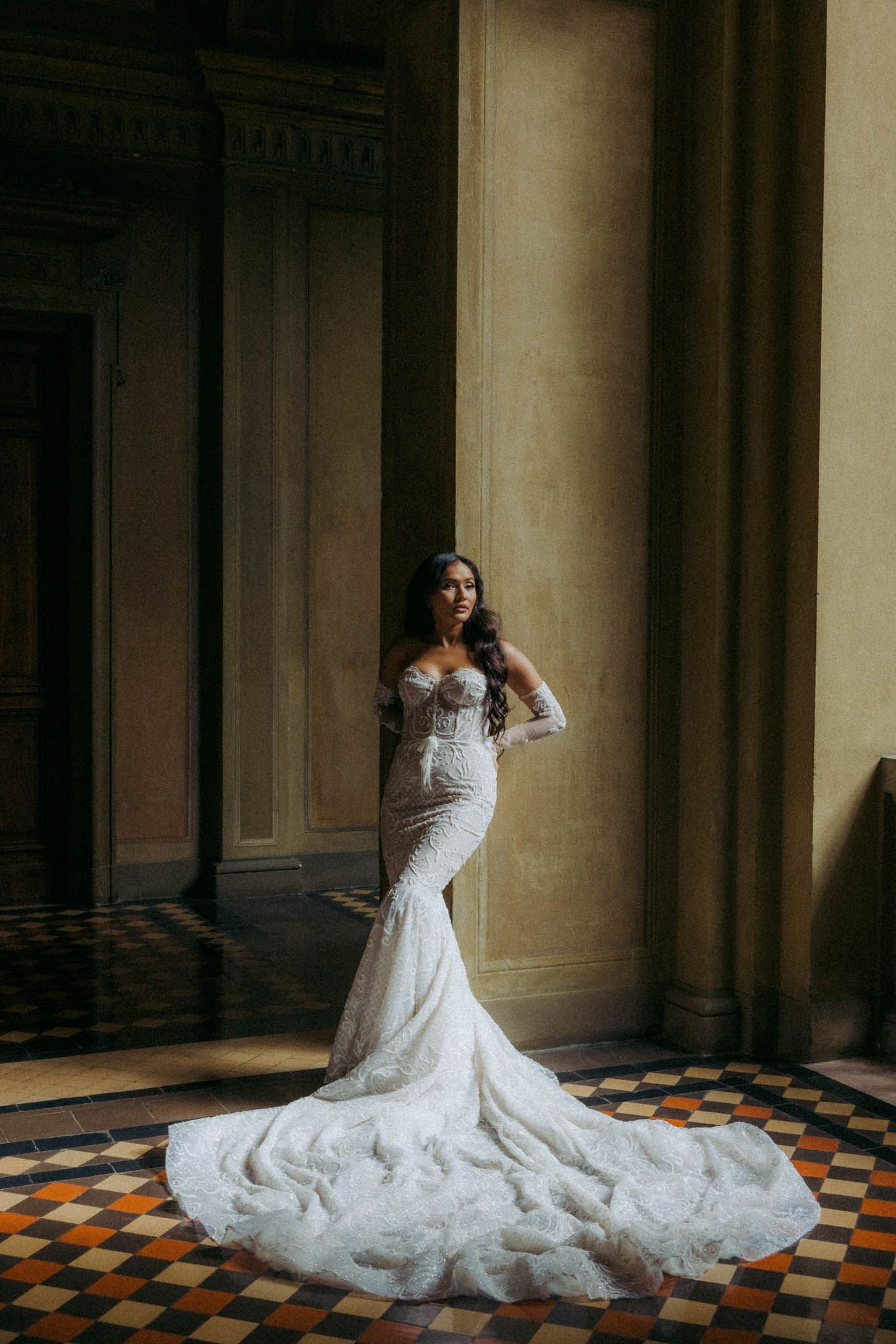 A bride in a fitted lace wedding gown with long sleeves and dramatic cathedral train poses in an elegant hallway.
