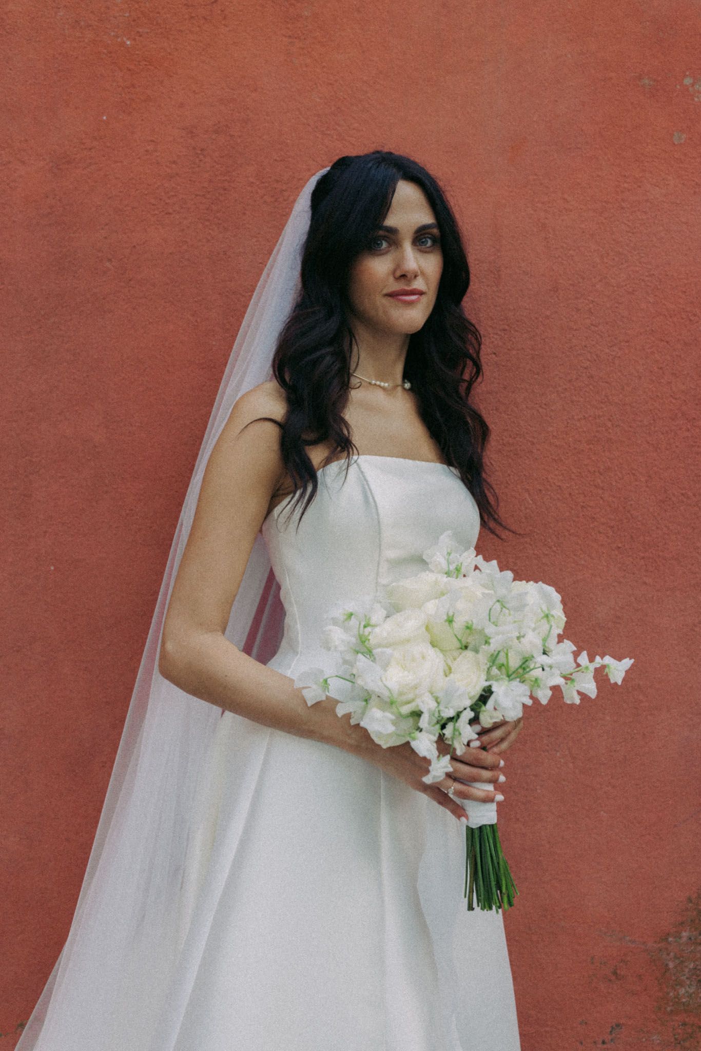 A bride in a classic white gown holds a bouquet of white flowers against a red wall backdrop.