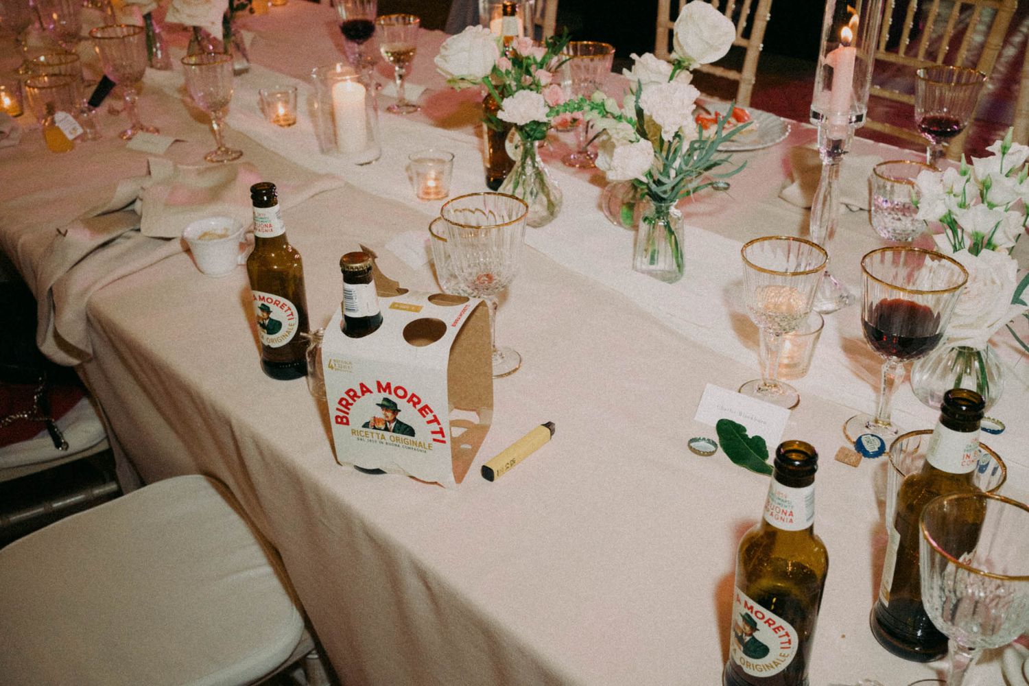 Wedding reception table decorated with wine bottles, floral centerpieces, and elegant place settings on white tablecloth.
