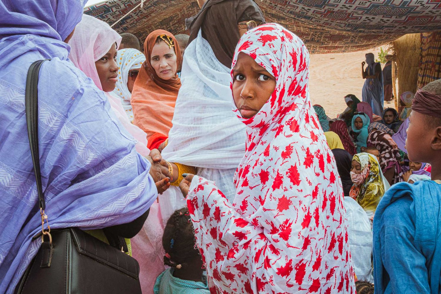 A gathering of people in colorful headscarves and traditional dress under a shaded outdoor space.