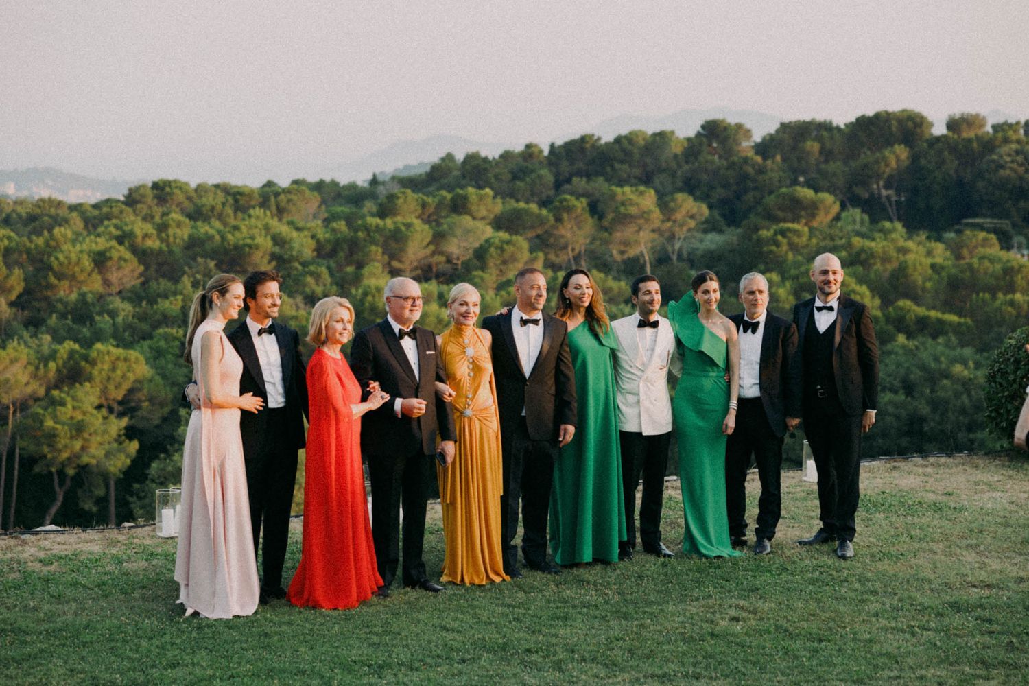 Wedding party in colorful rainbow-ordered dresses and black suits stands in a line on a grassy hillside at sunset.