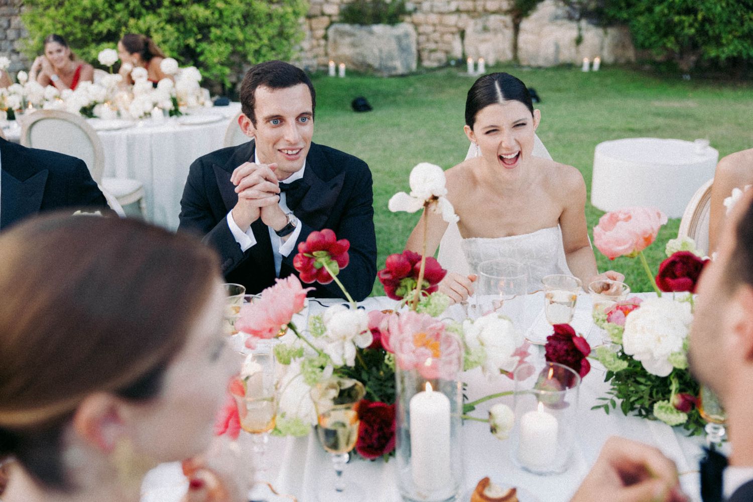 A couple shares a joyful moment at their outdoor wedding reception table decorated with pink and red floral centerpieces.