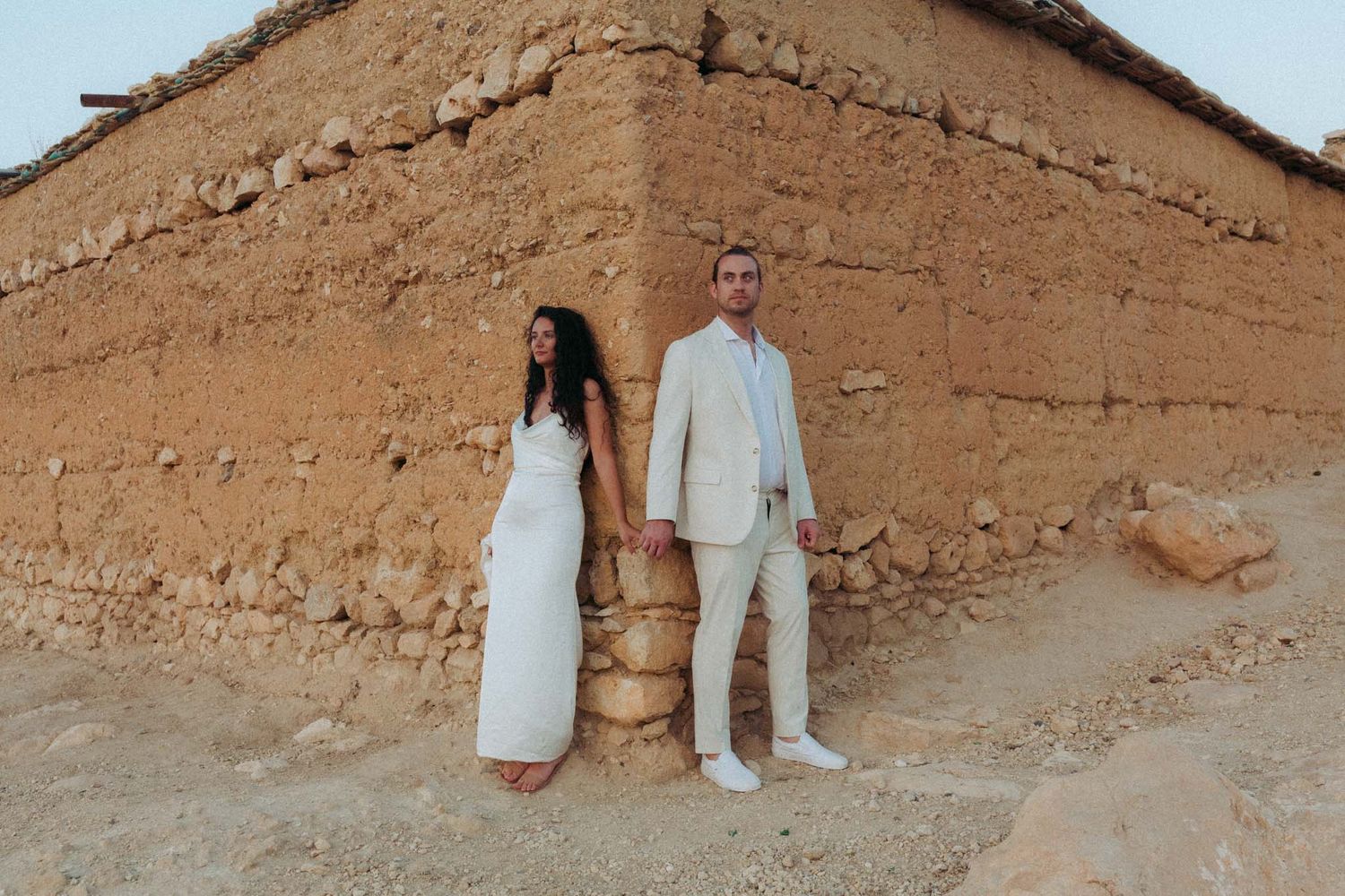 Two people in white attire stand against an earthen adobe wall in a desert setting.