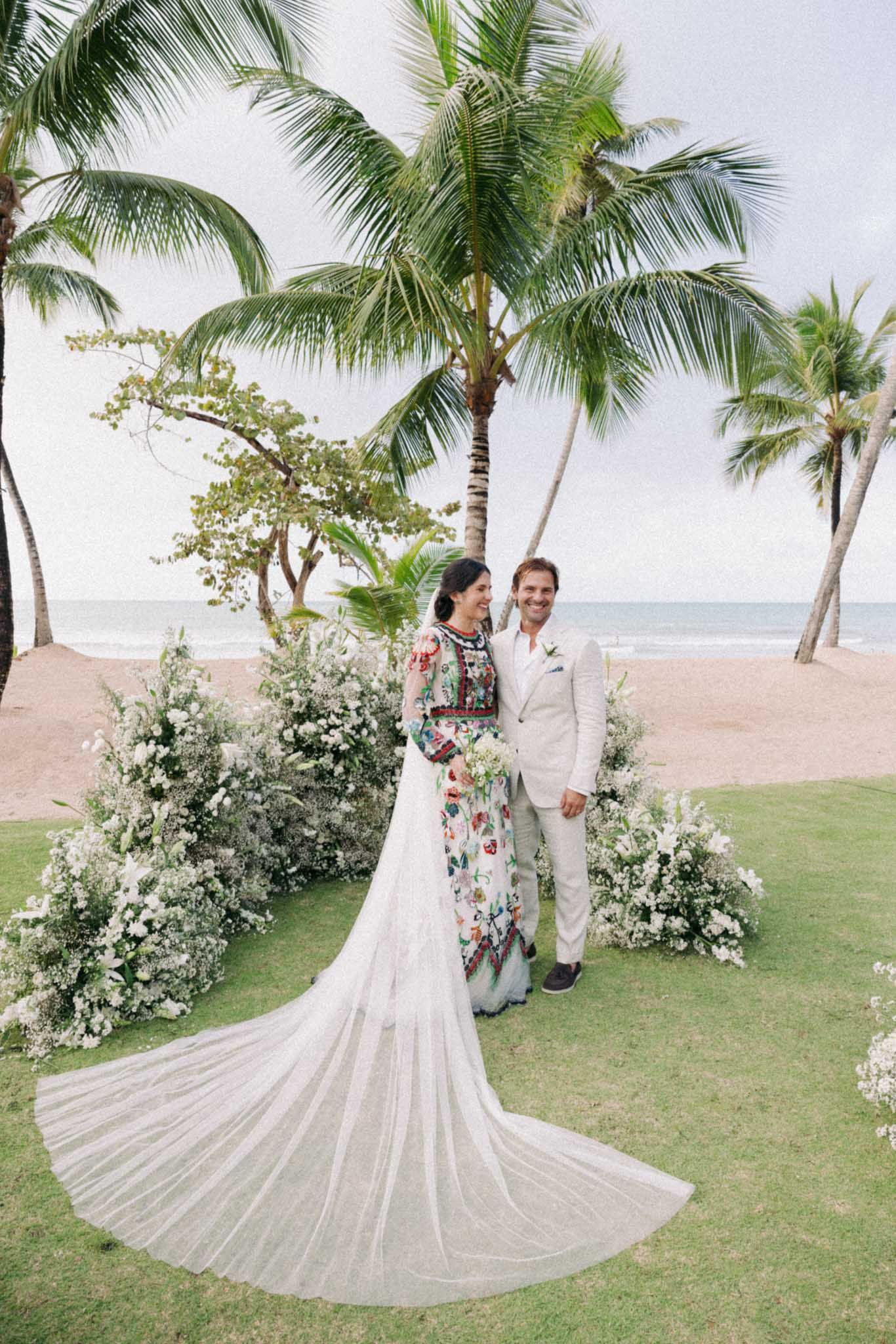 A tropical beach wedding scene with palm trees, white floral decorations, and a long-trained wedding dress on manicured grass.