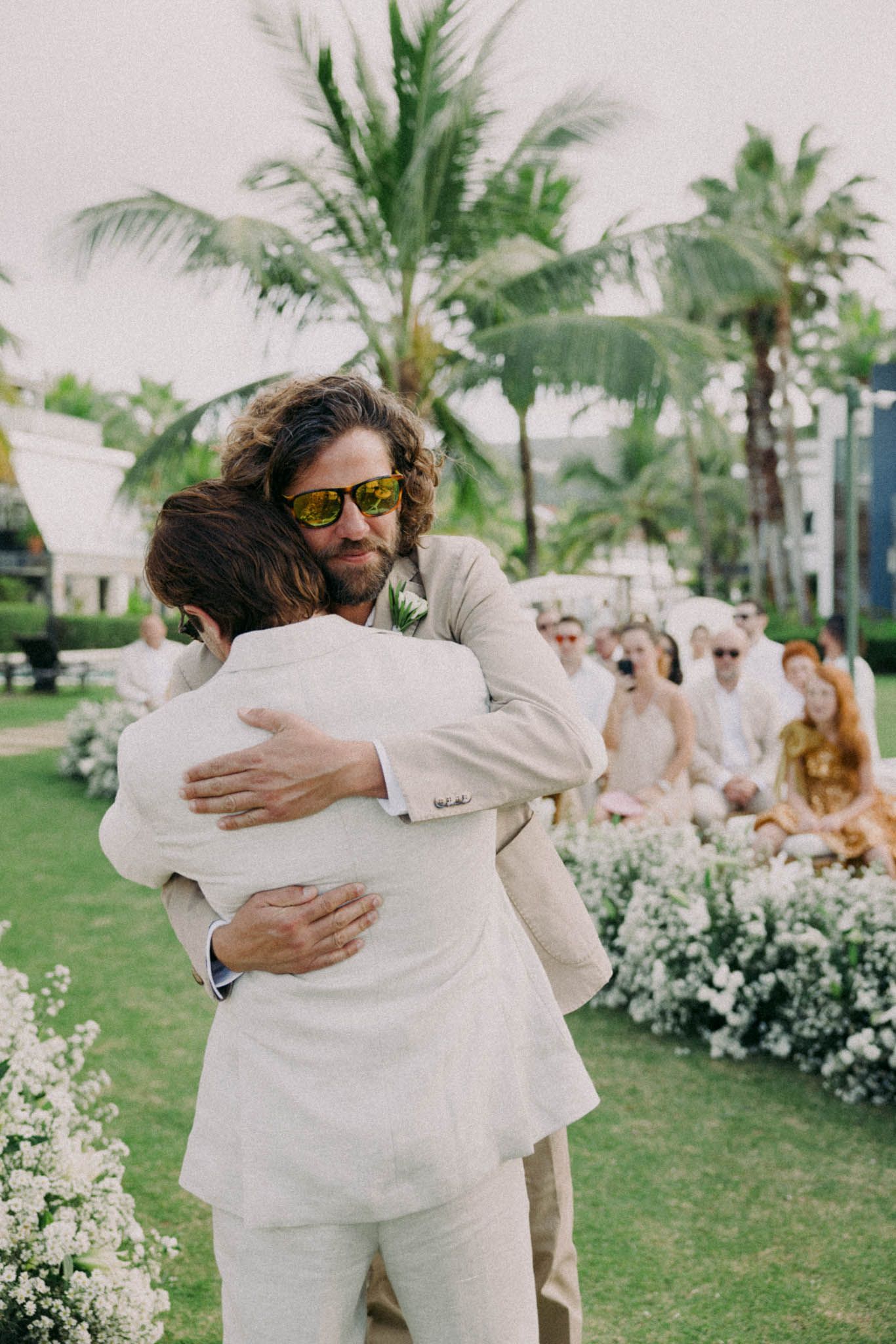 Two people in white attire share an emotional embrace on a lush green lawn with palm trees and white flowers.