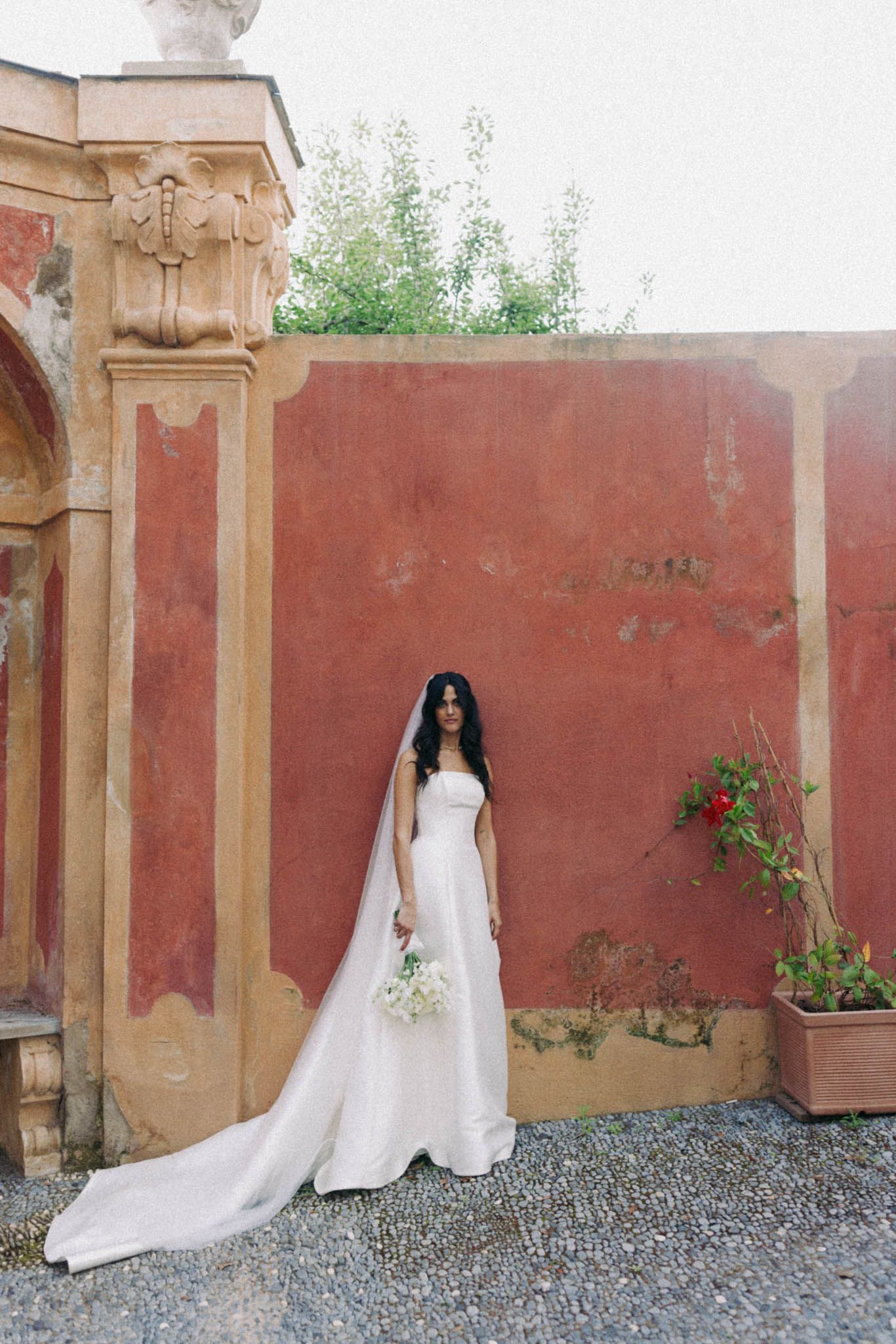 A bride in a white wedding gown with long train stands against a rustic terracotta wall with potted plants.