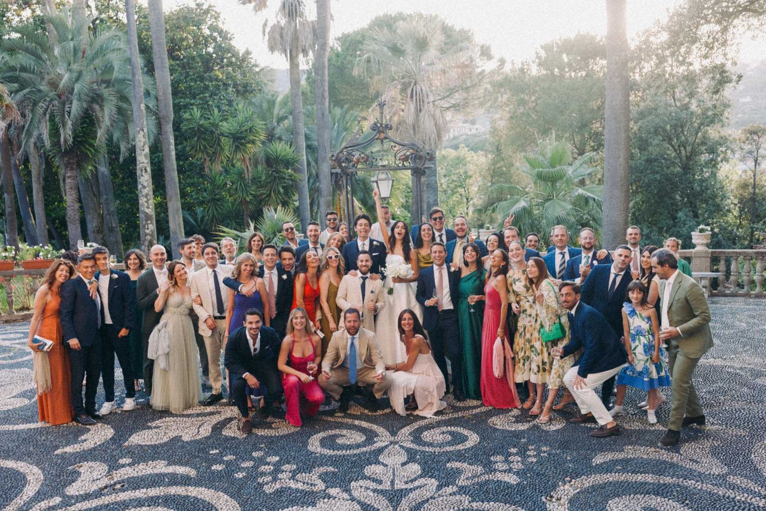 Large wedding party poses together in formal attire on outdoor garden path with palm trees and greenery in background.