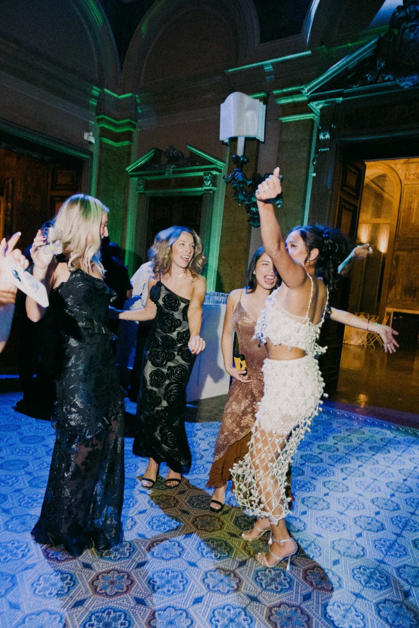 Party guests dance and celebrate on a blue patterned carpet under dramatic green lighting at an evening event.
