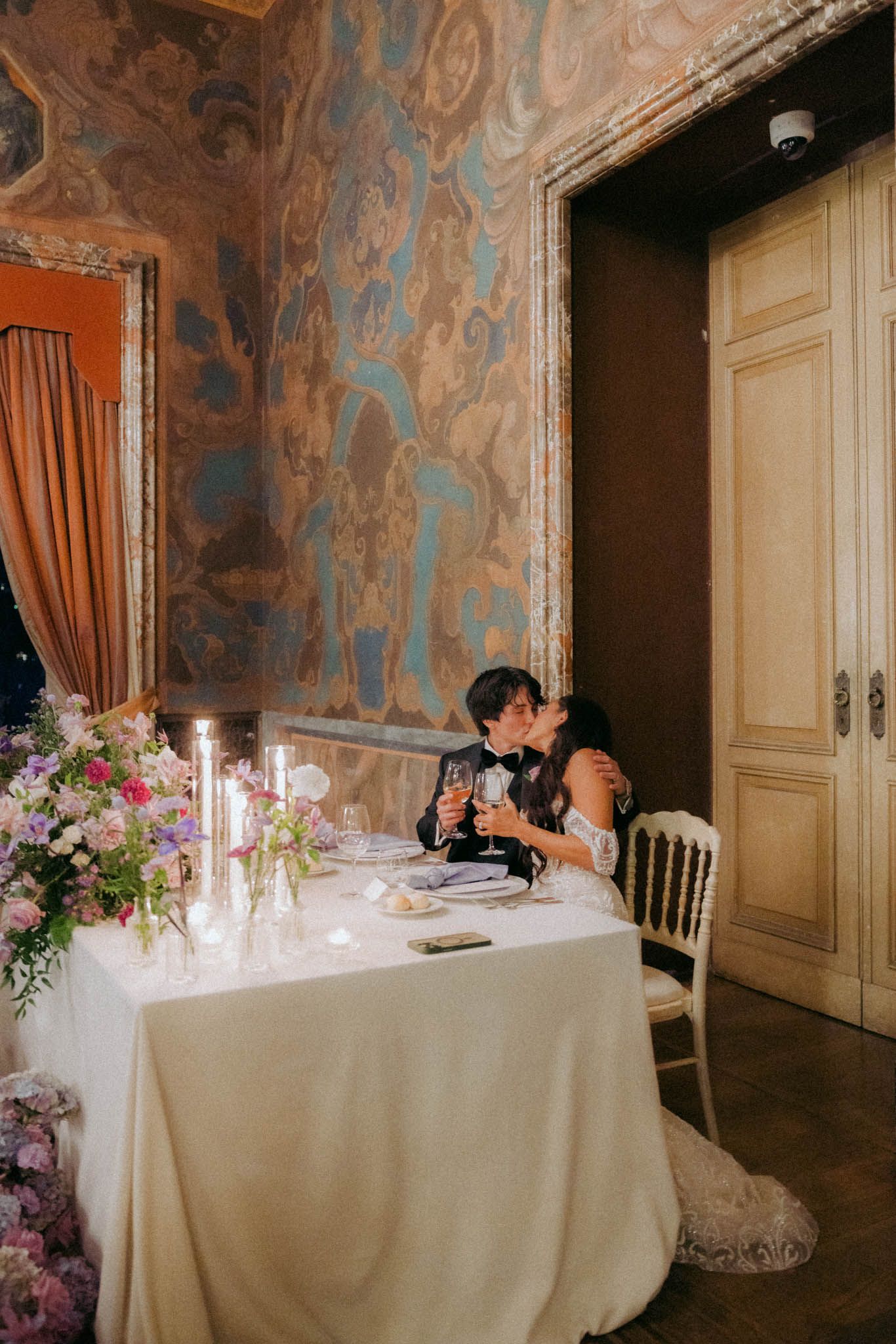 Elegant wedding reception table with white linens, candles and floral arrangements in an ornate room with damask wallpaper.