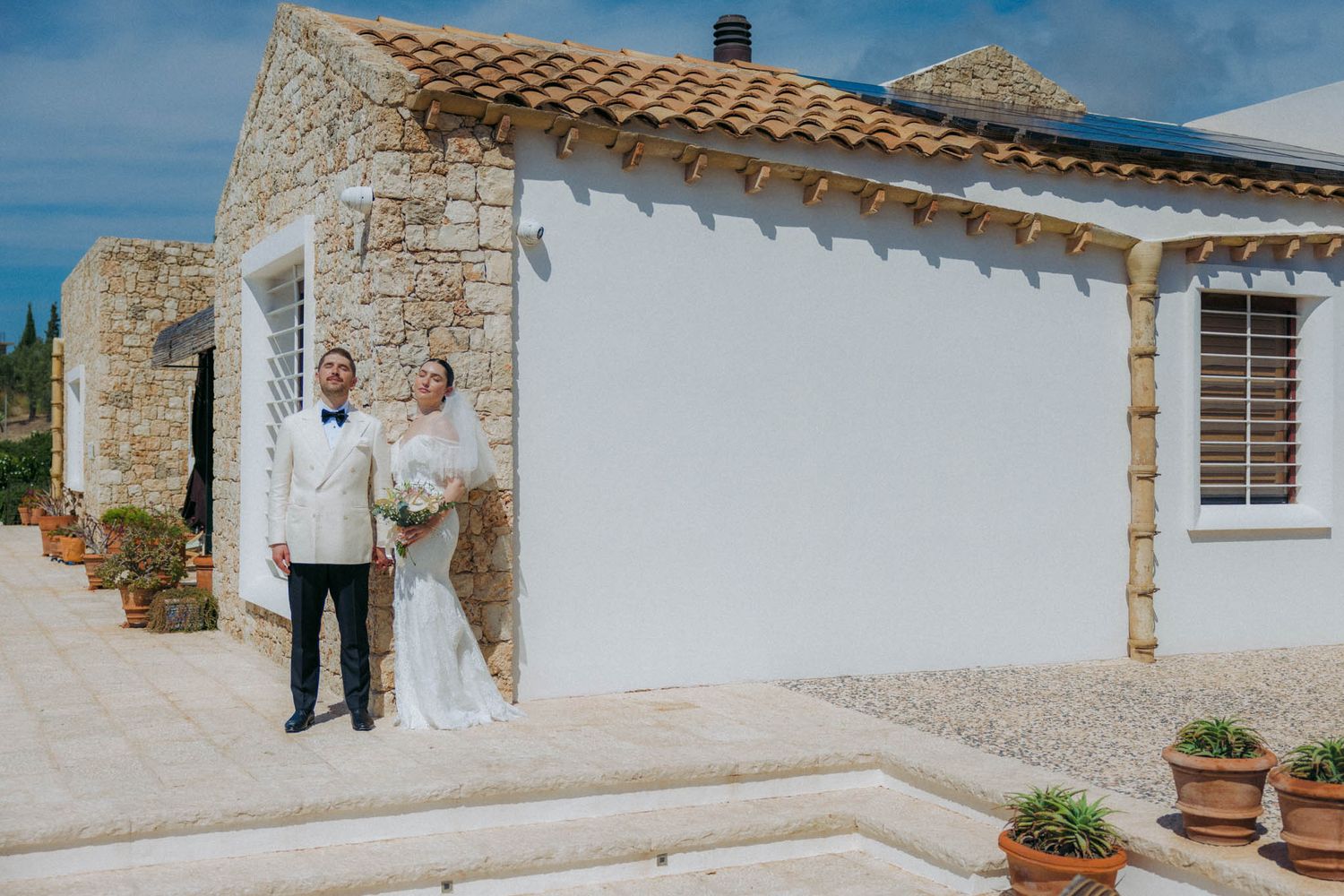 A series of white stucco buildings with terracotta roof tiles and stone walls under a bright blue Mediterranean sky.