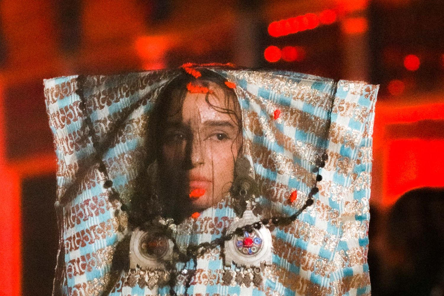 A performer on stage with a sheer patterned fabric draped over them in dramatic red lighting.