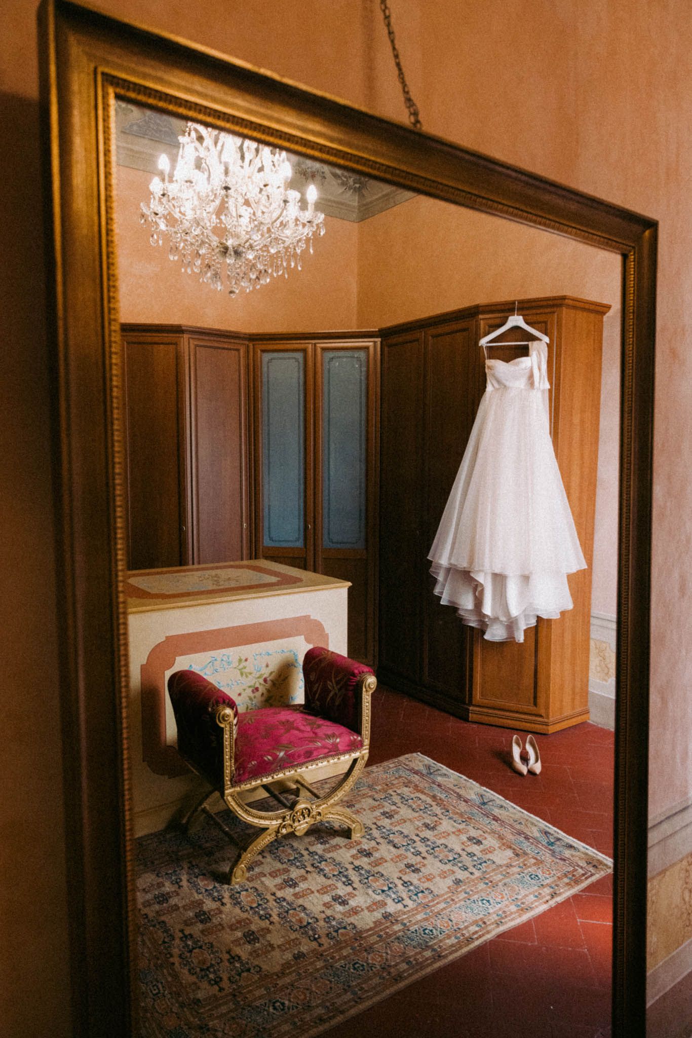 Elegant white wedding dress hangs in a luxurious hotel room with crystal chandeliers and antique furniture.