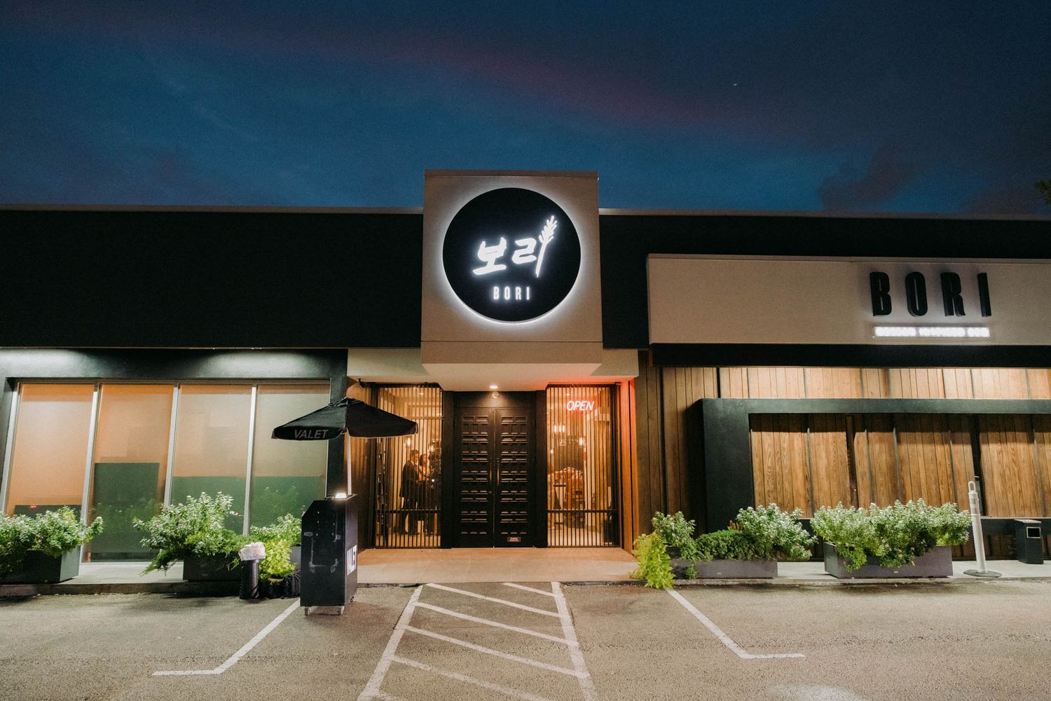 Modern restaurant storefront with illuminated logo and entrance at night, featuring planters and clean architectural lines.