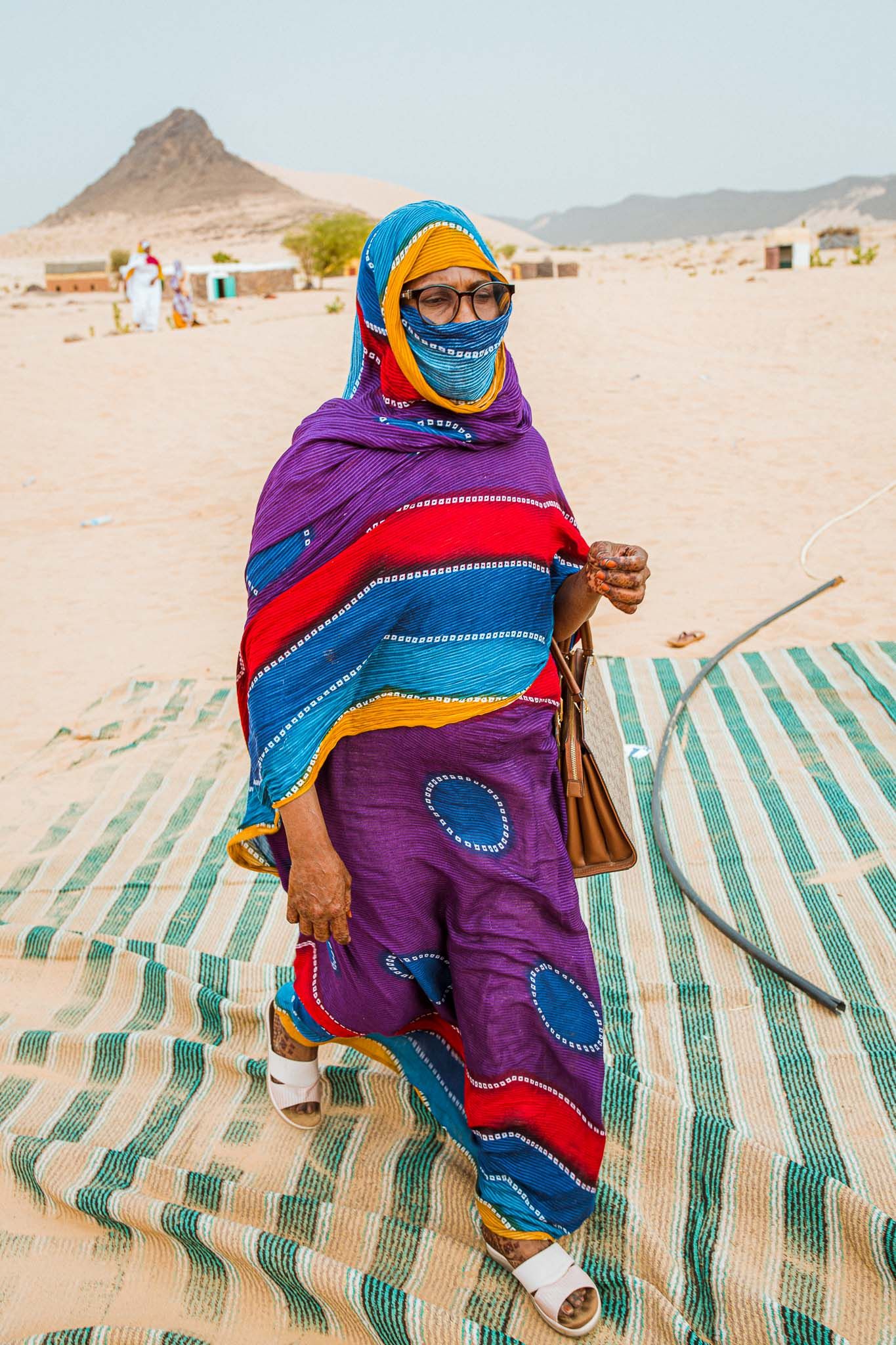 Person wearing colorful traditional dress with purple and blue patterns standing on desert sand.