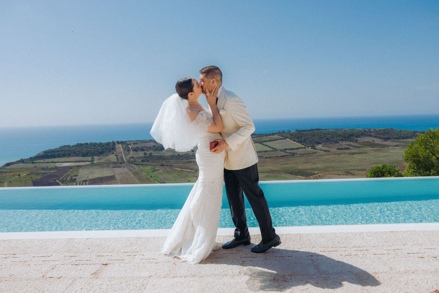 Newlyweds embrace on a scenic coastal cliff overlooking turquoise waters and rolling hills in the distance.