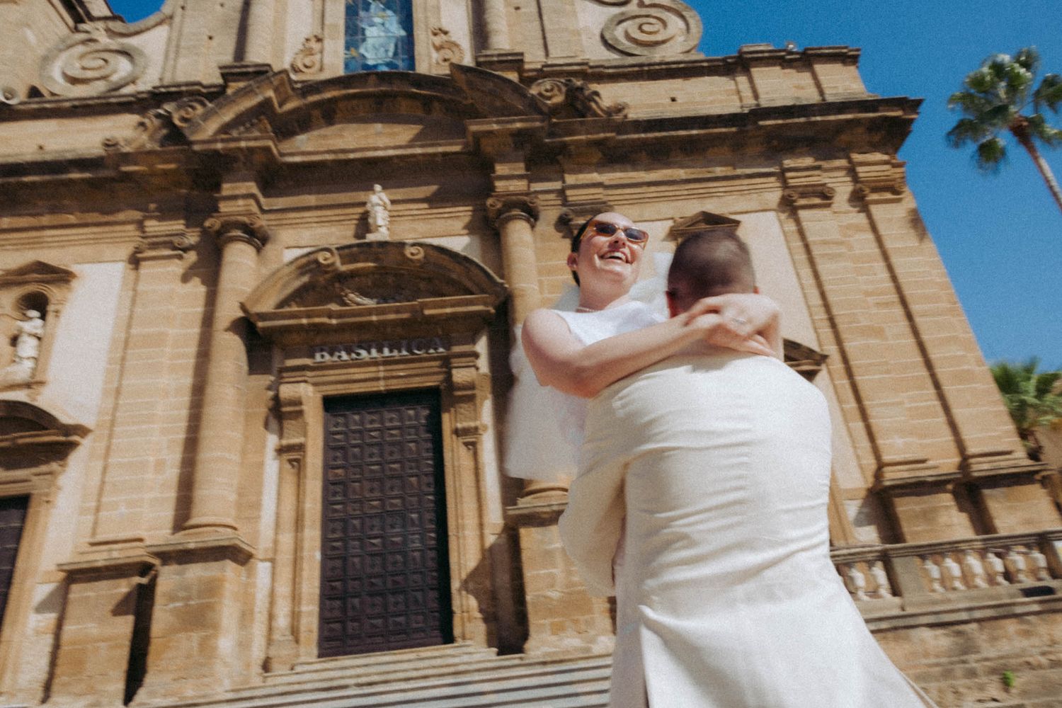 Two people in white embrace in front of an ornate stone church facade with a palm tree against a bright blue sky.