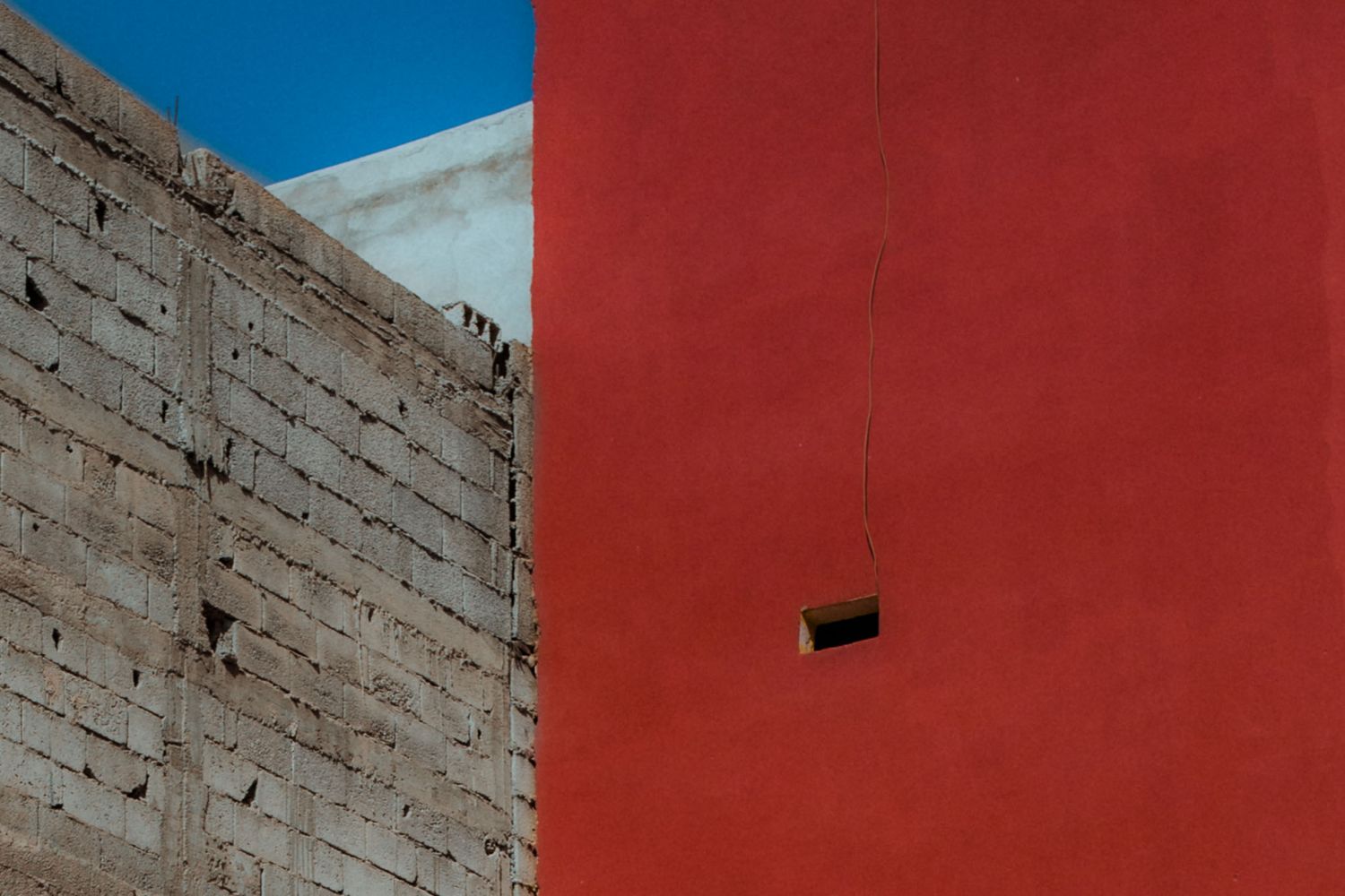Architectural detail showing a textured gray brick wall against a vibrant red painted surface under blue sky.