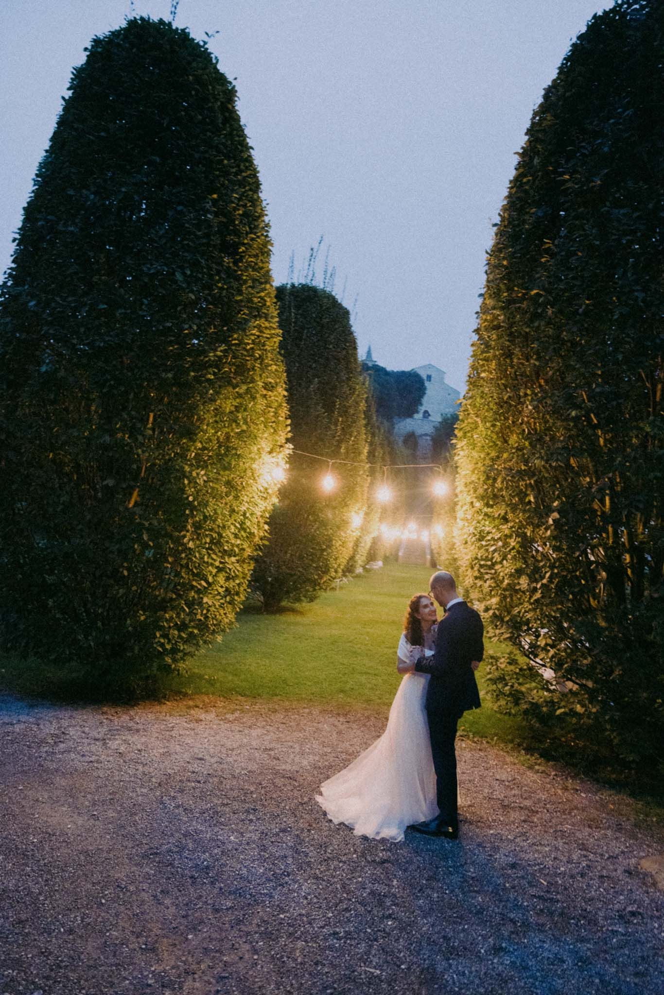 A romantic evening photo series showing a couple embracing on a path lined with illuminated topiary trees.