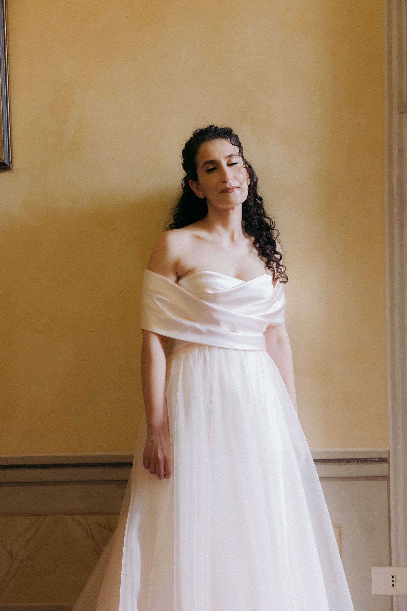 A person in a flowing white off-shoulder dress poses gracefully against a beige wall in a series of artistic shots.