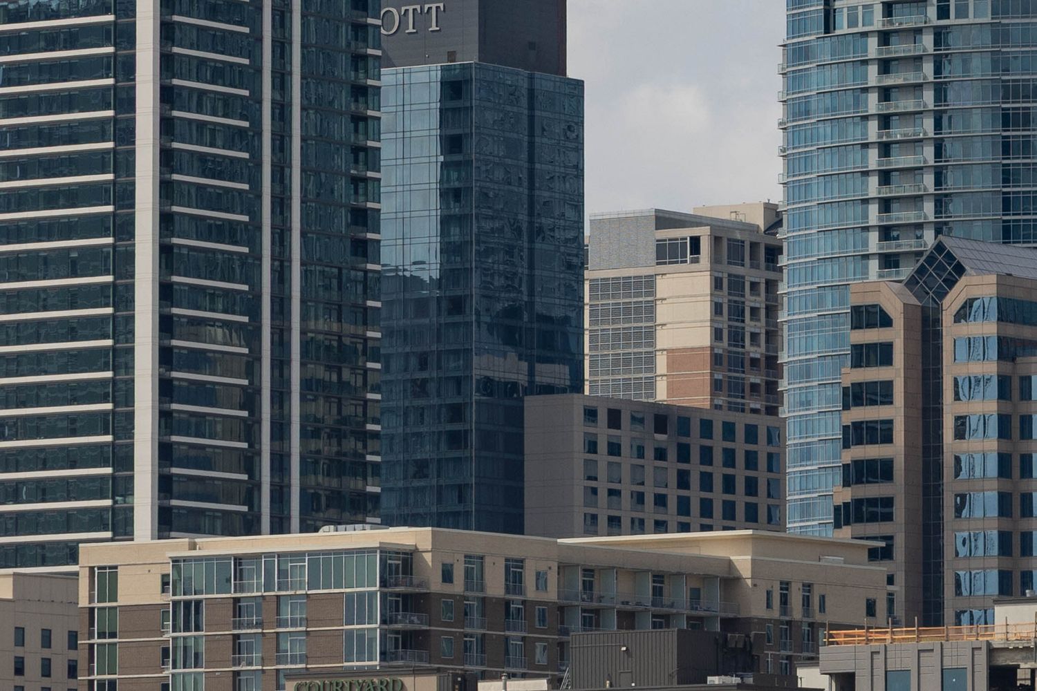 Modern glass skyscrapers and high-rise apartment buildings line an urban waterfront on a cloudy day.