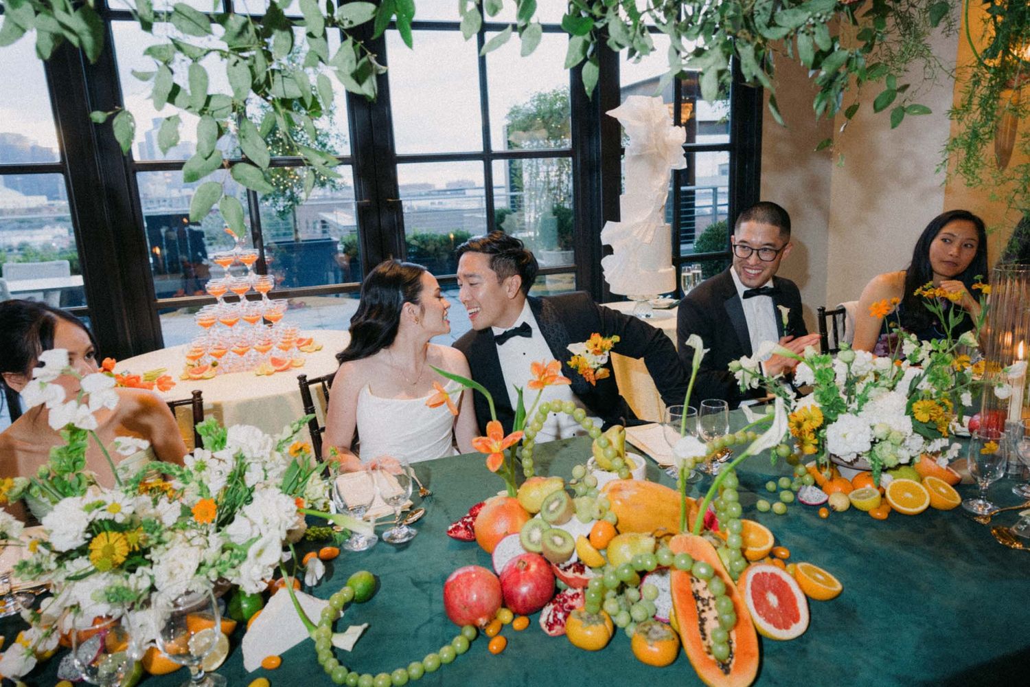 Elegant wedding reception table decorated with fresh fruits and flowers in a bright venue with large windows.