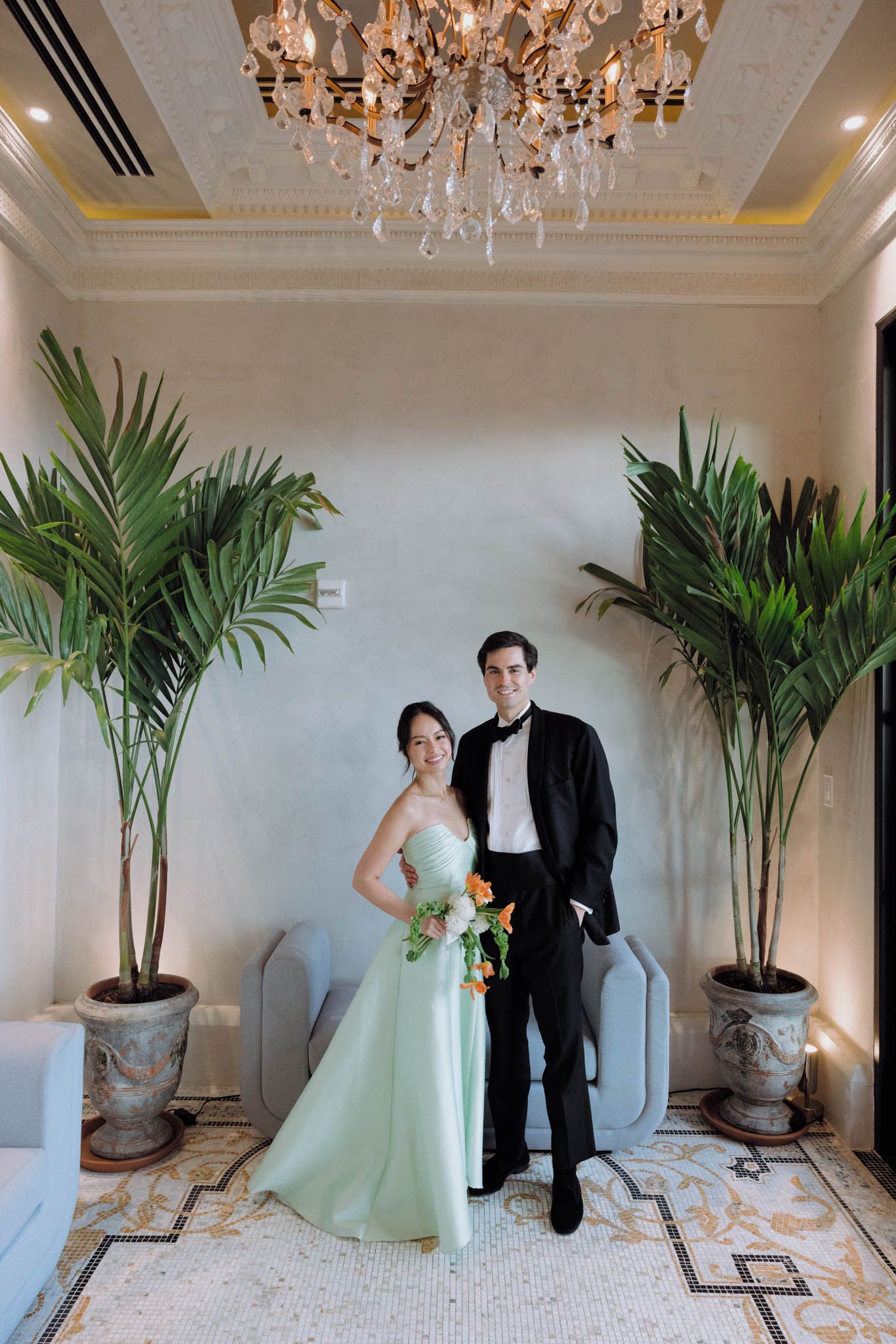 A couple poses together in an elegant room with potted palm plants and a crystal chandelier overhead.