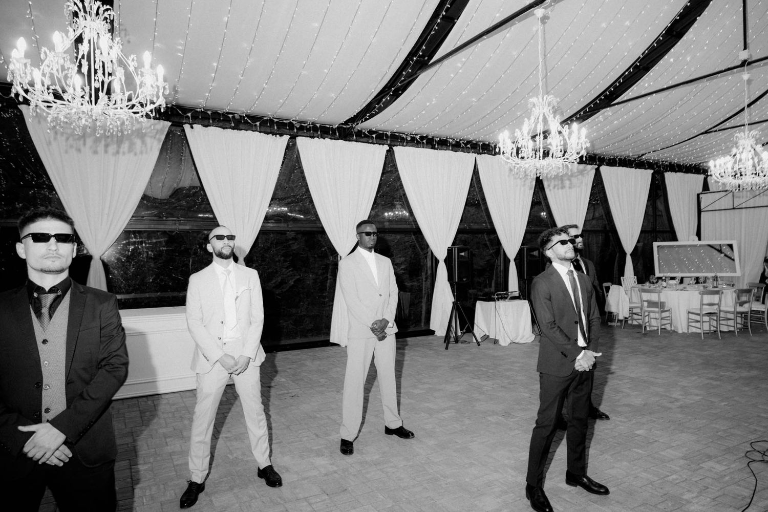 Black and white photo of a group in sunglasses and suits standing in an elegant event space with crystal chandeliers.