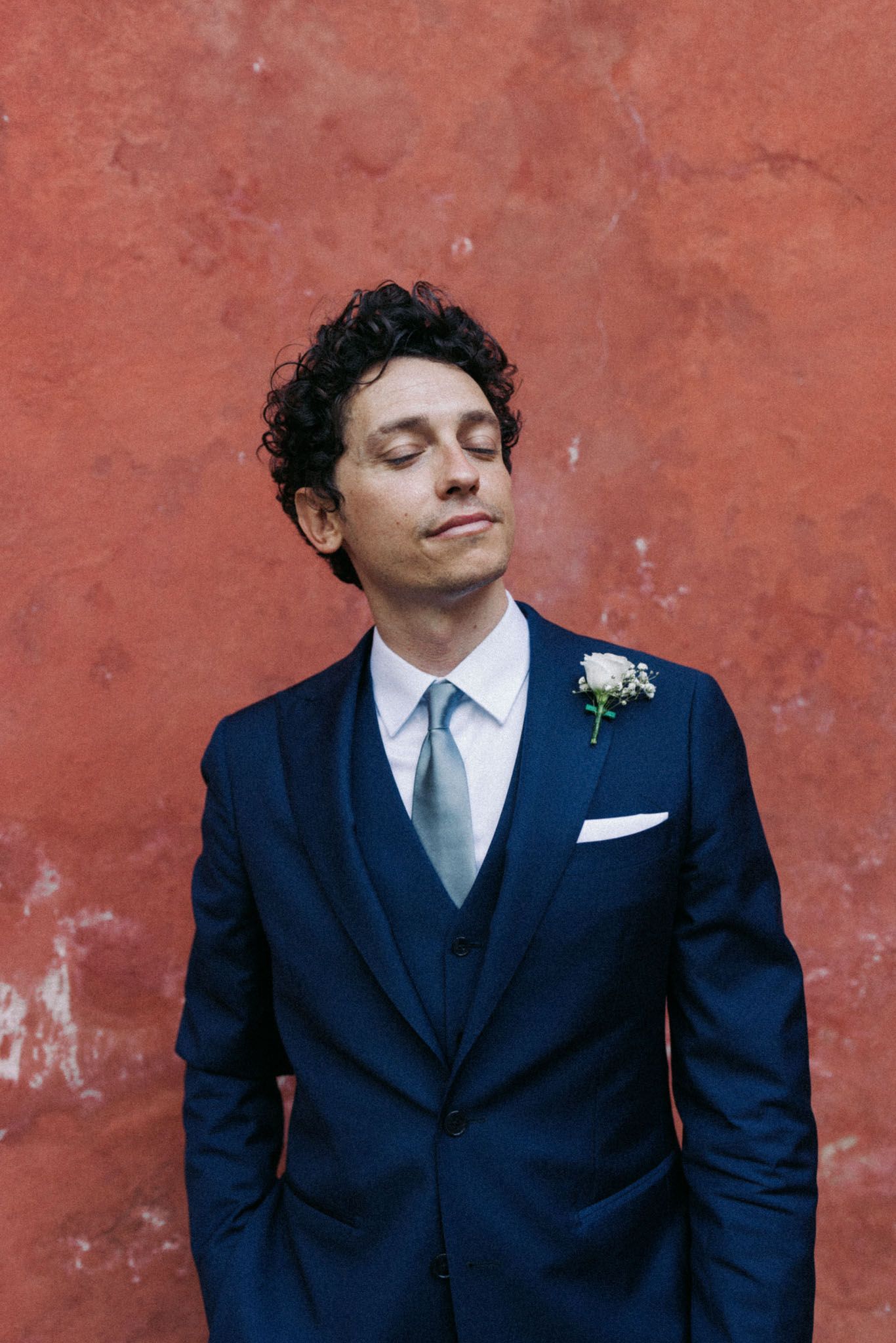 Person in navy blue suit with curly hair smiles against rustic red wall in sequence of four similar poses.