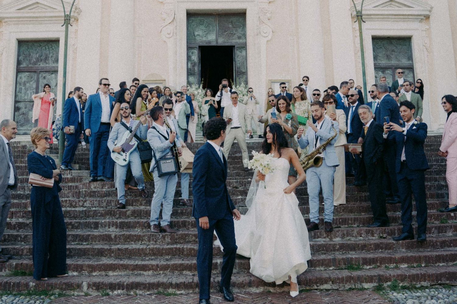 Wedding guests gather on stone steps outside a historic building during a formal celebration.