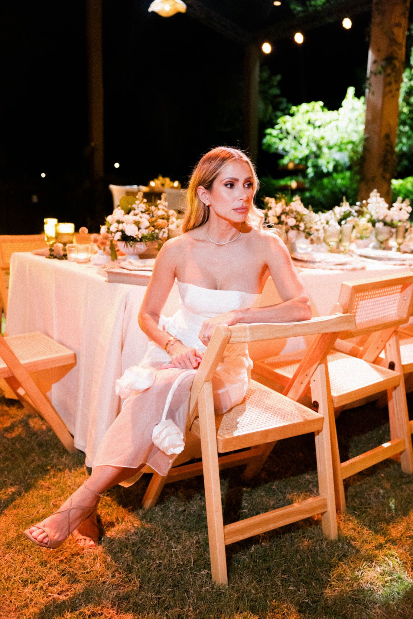A woman in a white dress sits at an elegant outdoor dinner table decorated with flowers and string lights.