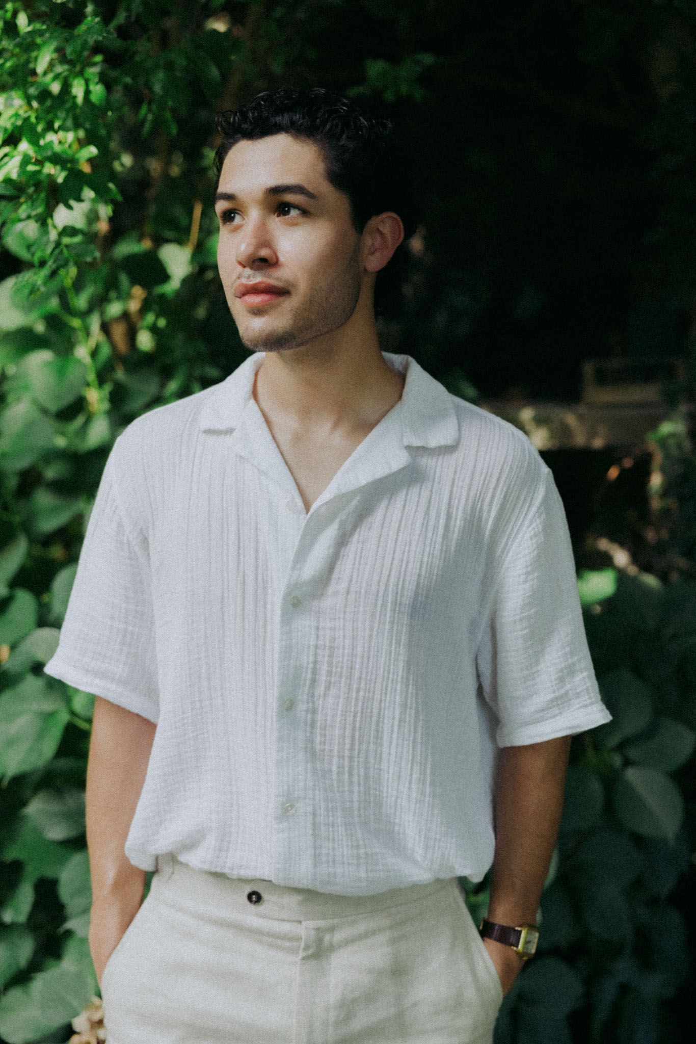 Person wearing a white short-sleeve button-down shirt and light colored pants poses outdoors against green foliage.