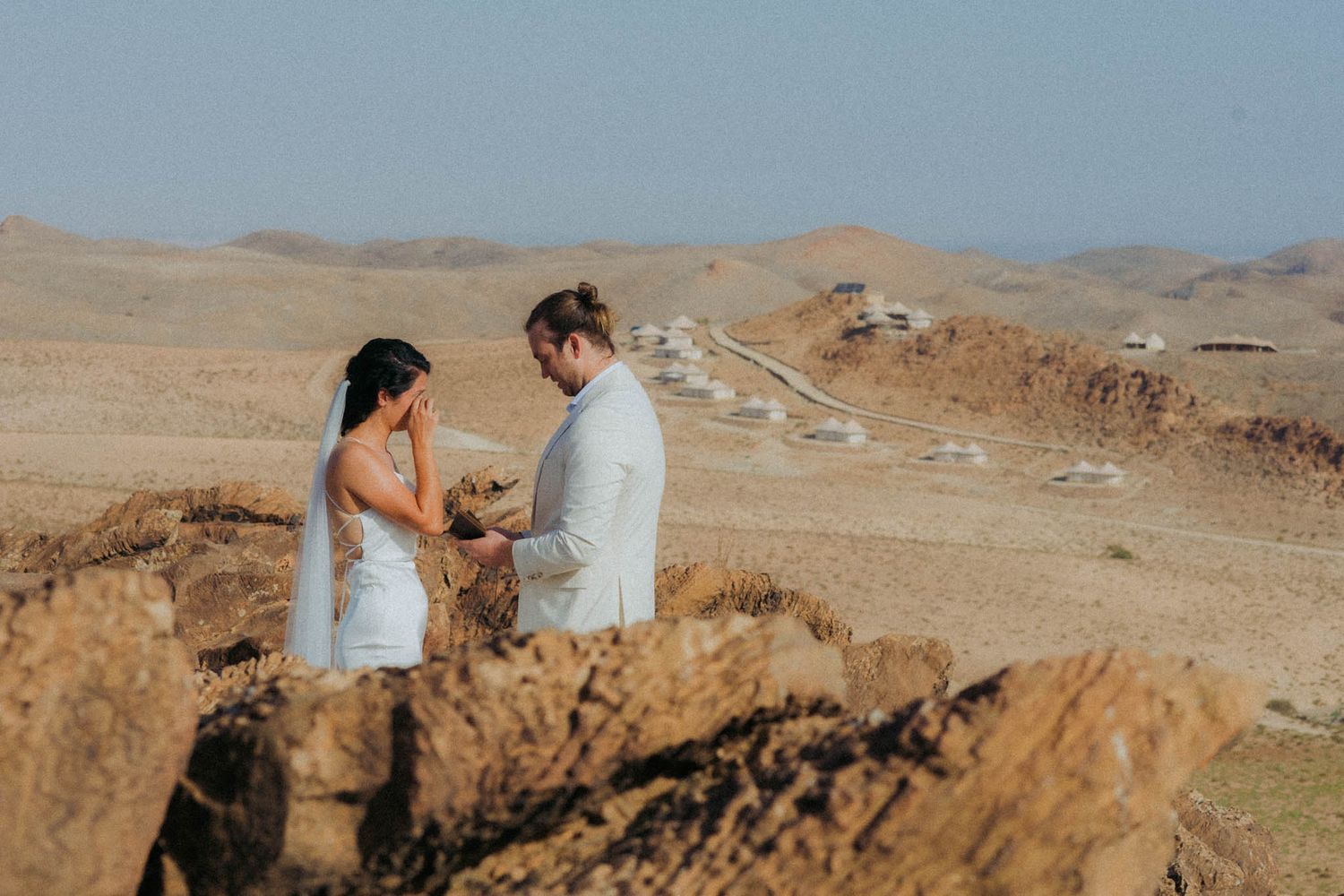 Group of hikers traverse rugged desert terrain with backpacks against backdrop of rocky mountains and blue sky.