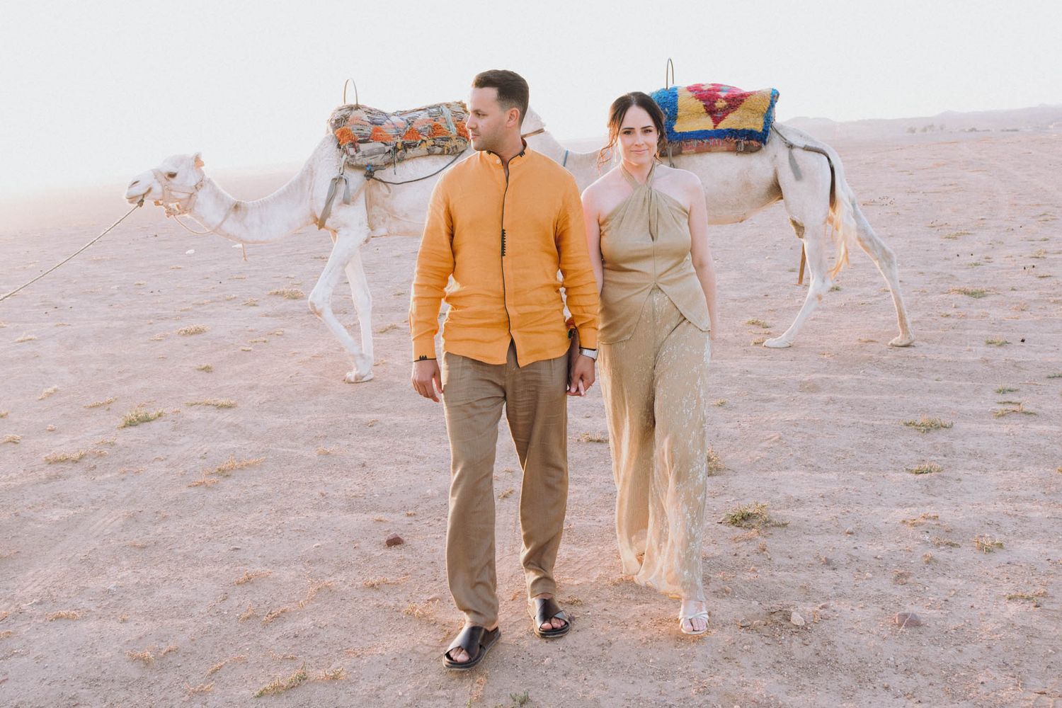 Couple in desert attire walking together with decorated camel in background against sandy landscape.