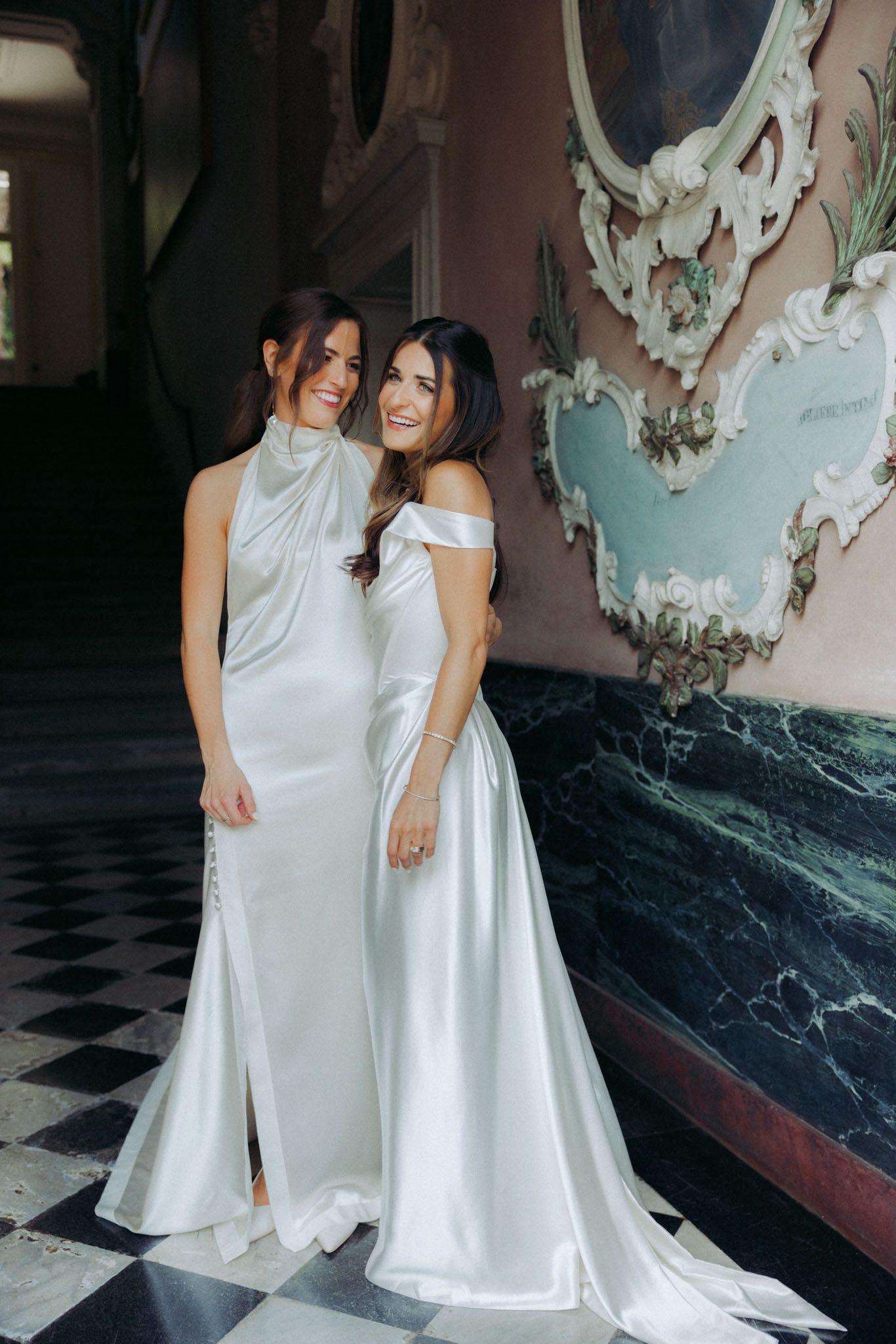 Two people in elegant white dresses stand together in an ornate room with a checkered floor and decorative walls.