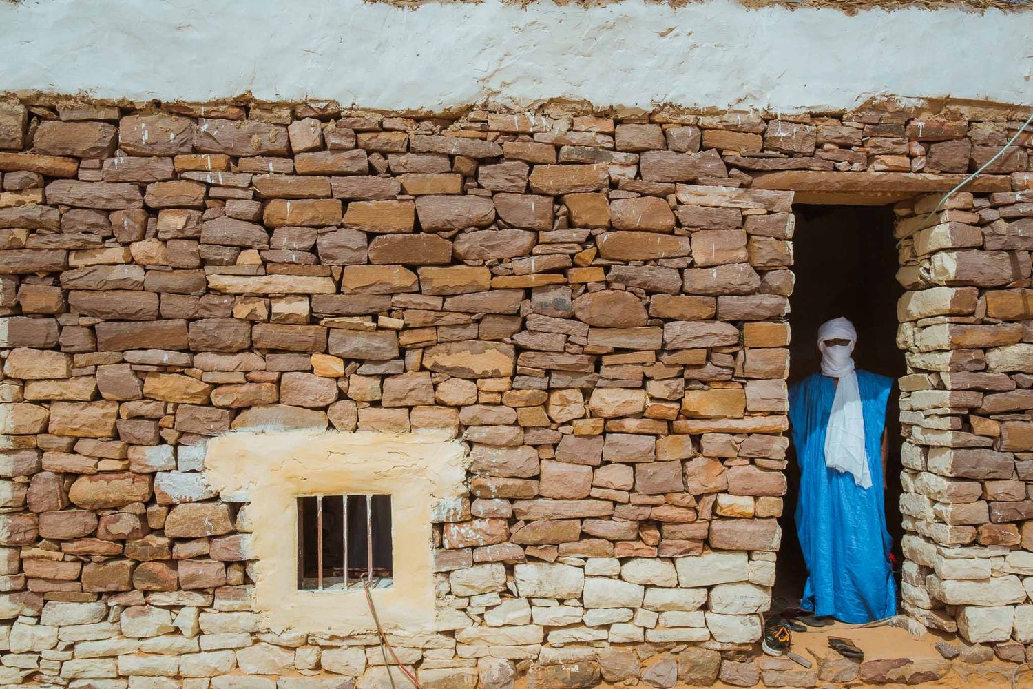 A figure in traditional blue robes stands in a doorway of a rustic stone building in a desert setting.