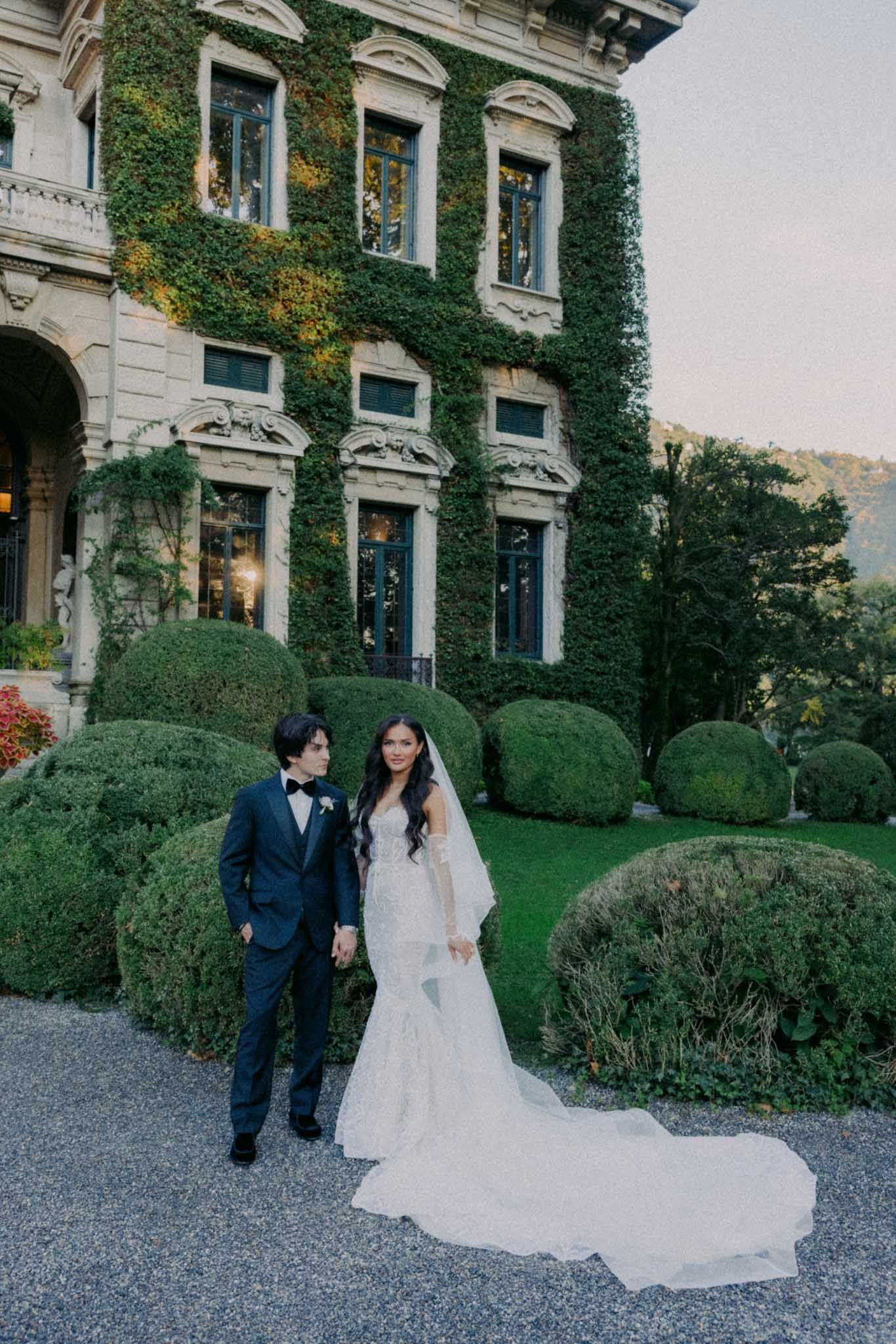 A couple poses for wedding photos in front of an elegant ivy-covered building with classic architecture.