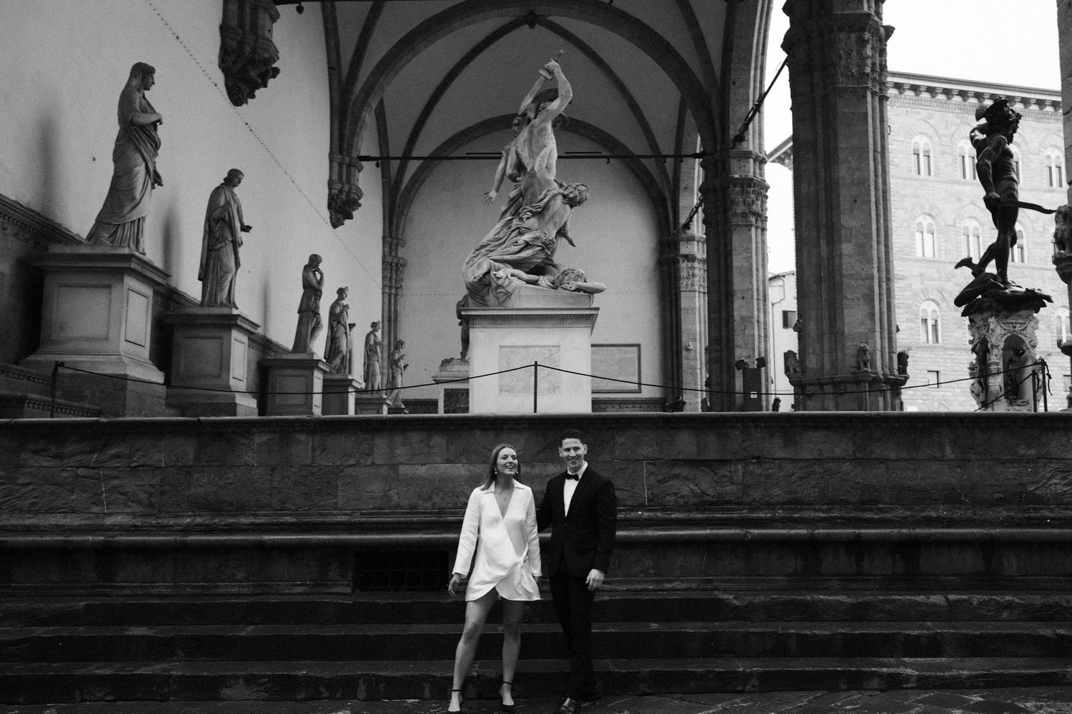 Artistic black and white photograph of couple walking past classical statues.
