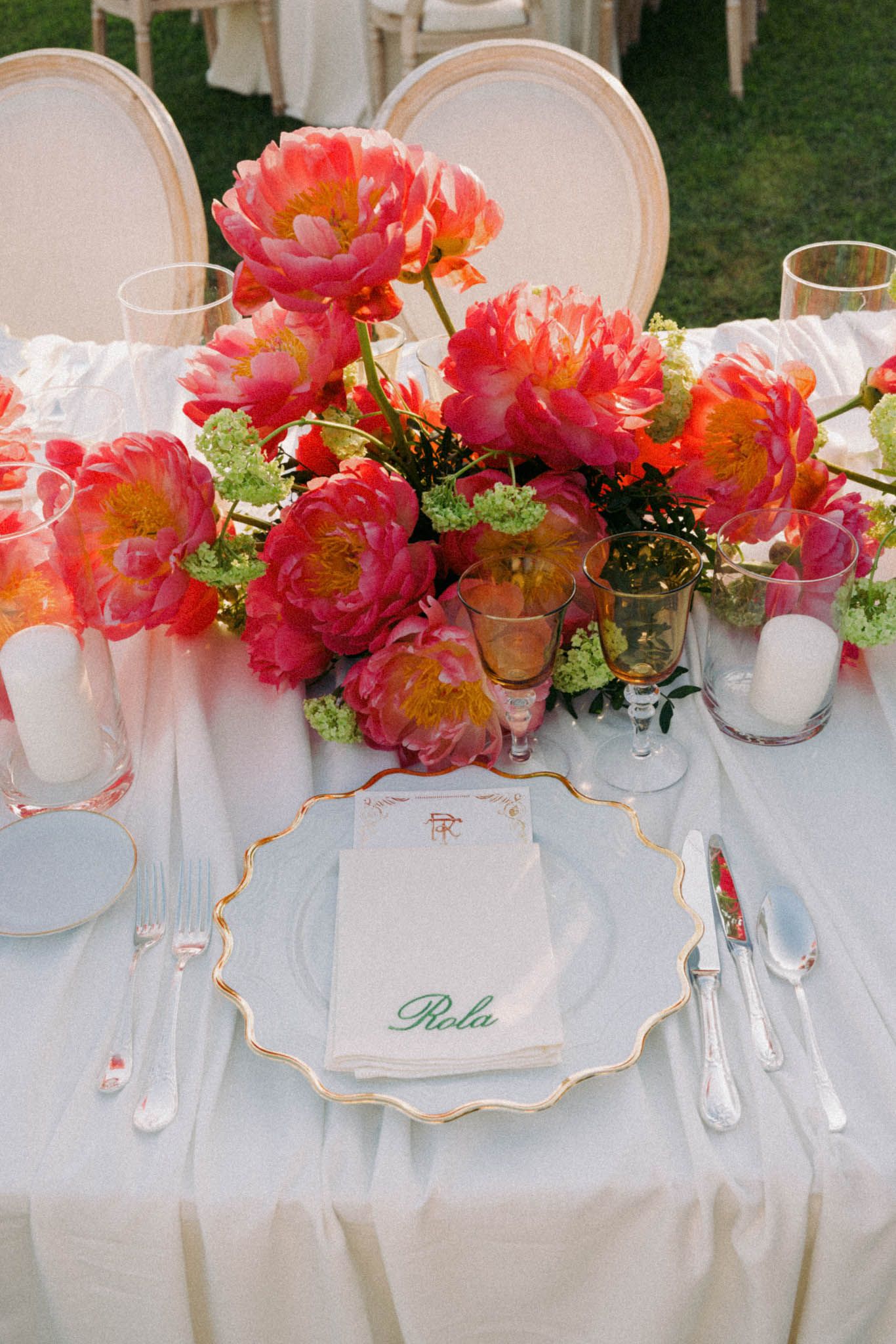 Elegant place setting with gold-rimmed plate and vibrant coral peonies centerpiece on white tablecloth at outdoor event.