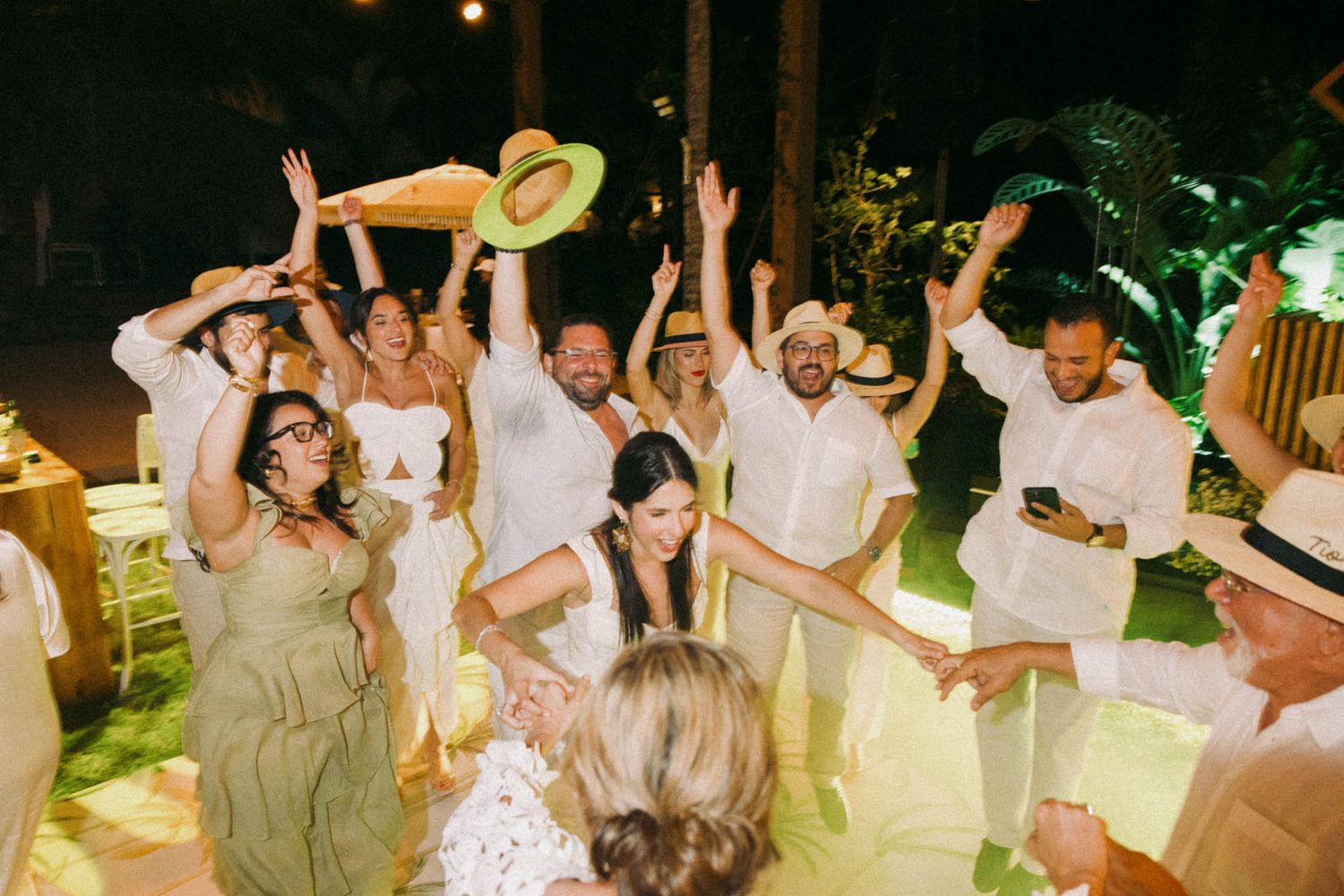 Wedding reception guests dancing and celebrating with raised arms on a festively lit outdoor dance floor.