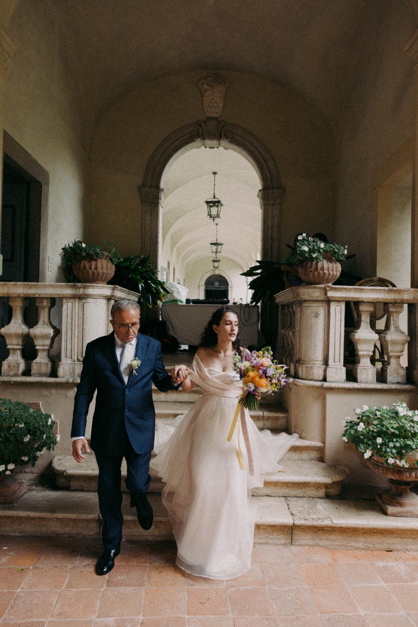 Couple walks through an elegant arched colonnade with terracotta tiles and potted plants.