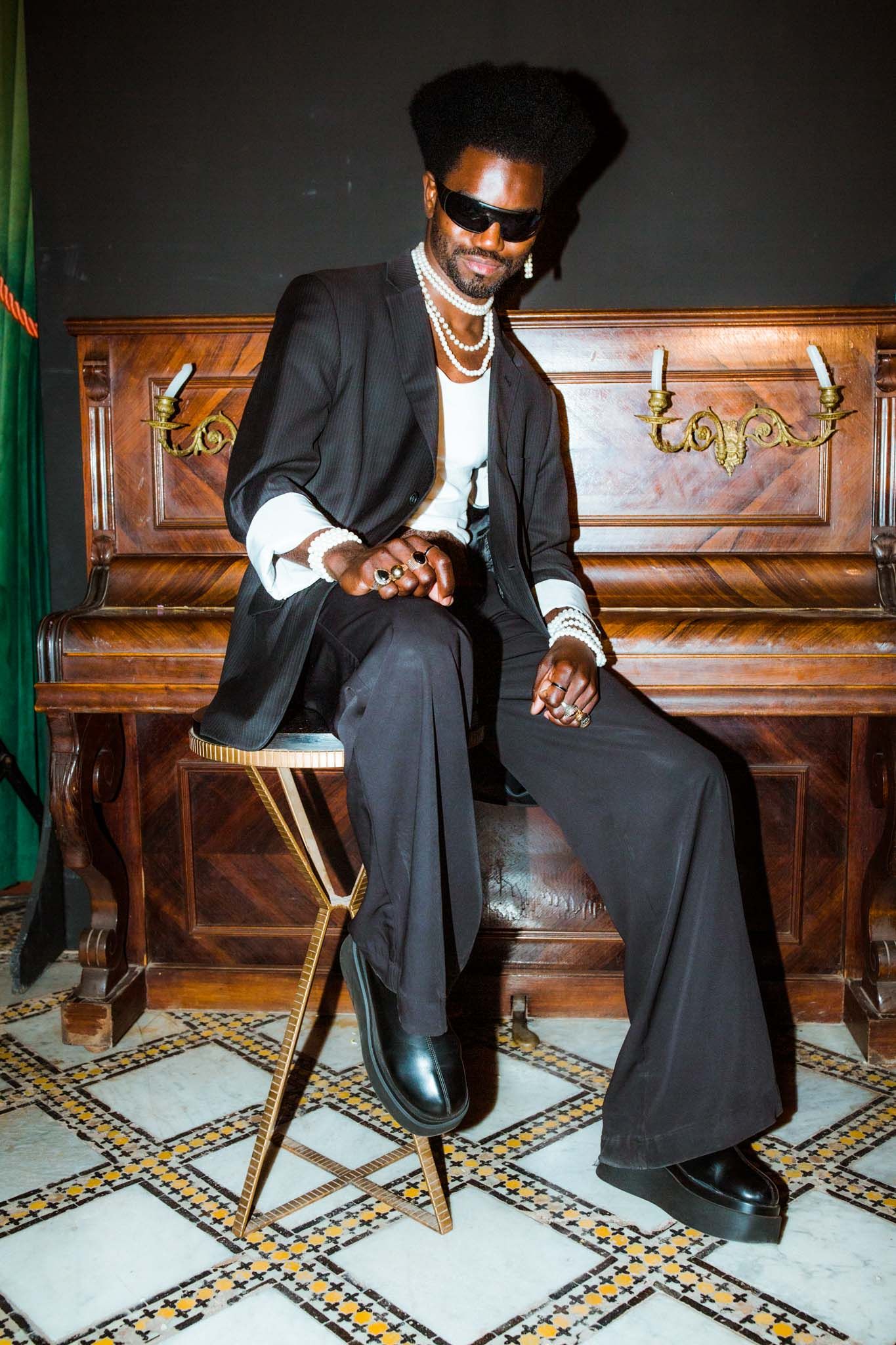 A sequence of photos showing someone in a black suit and sunglasses sitting on a wooden chair against a tiled floor.