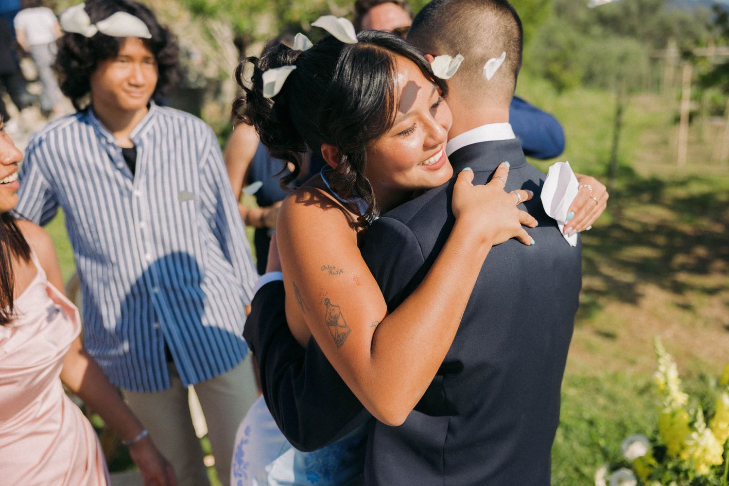 A couple shares a tender embrace while dancing together at an outdoor wedding celebration in a sunny garden setting.