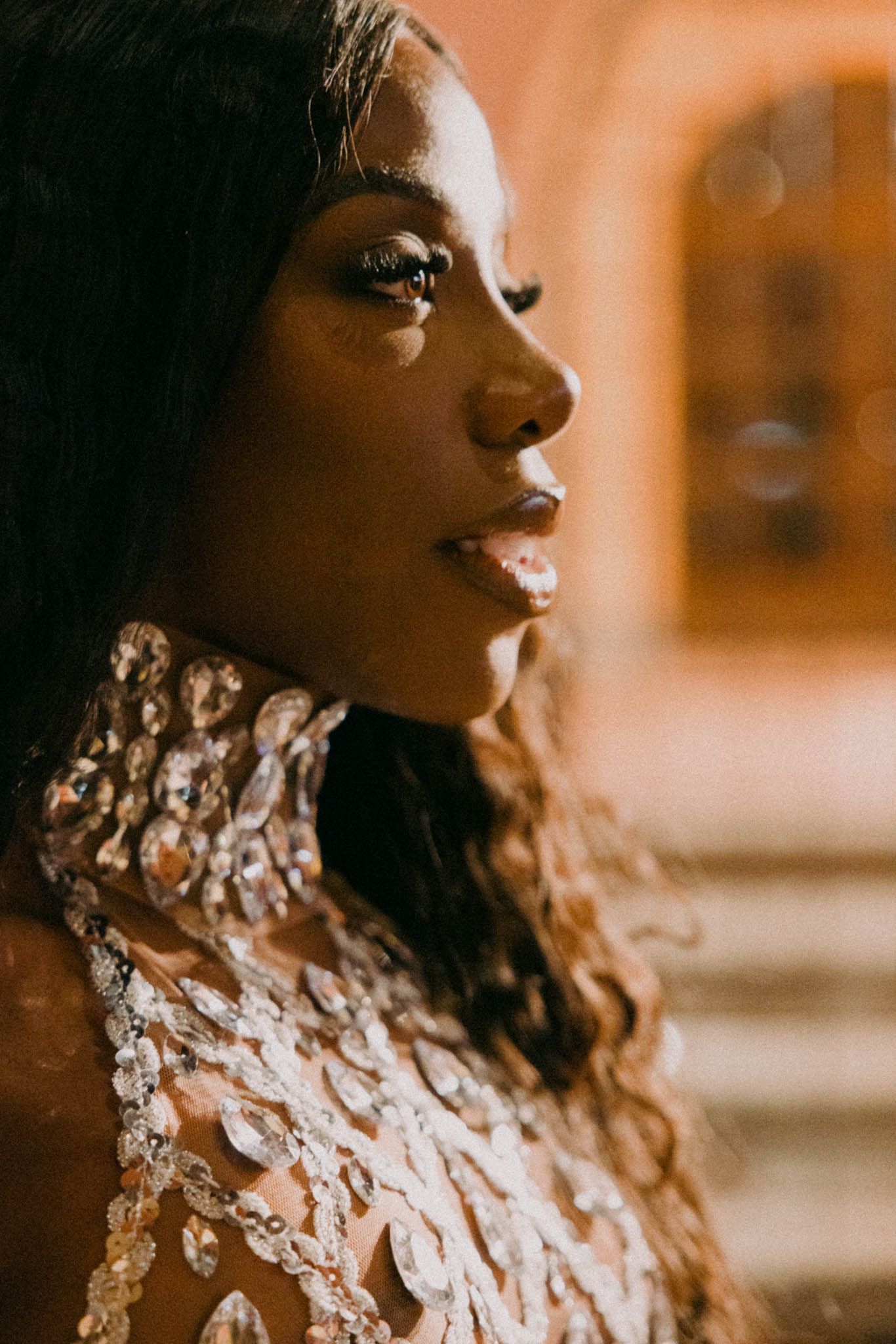 Side profile portrait in warm lighting showing a person wearing an ornate white lace or sequined high-neck dress.