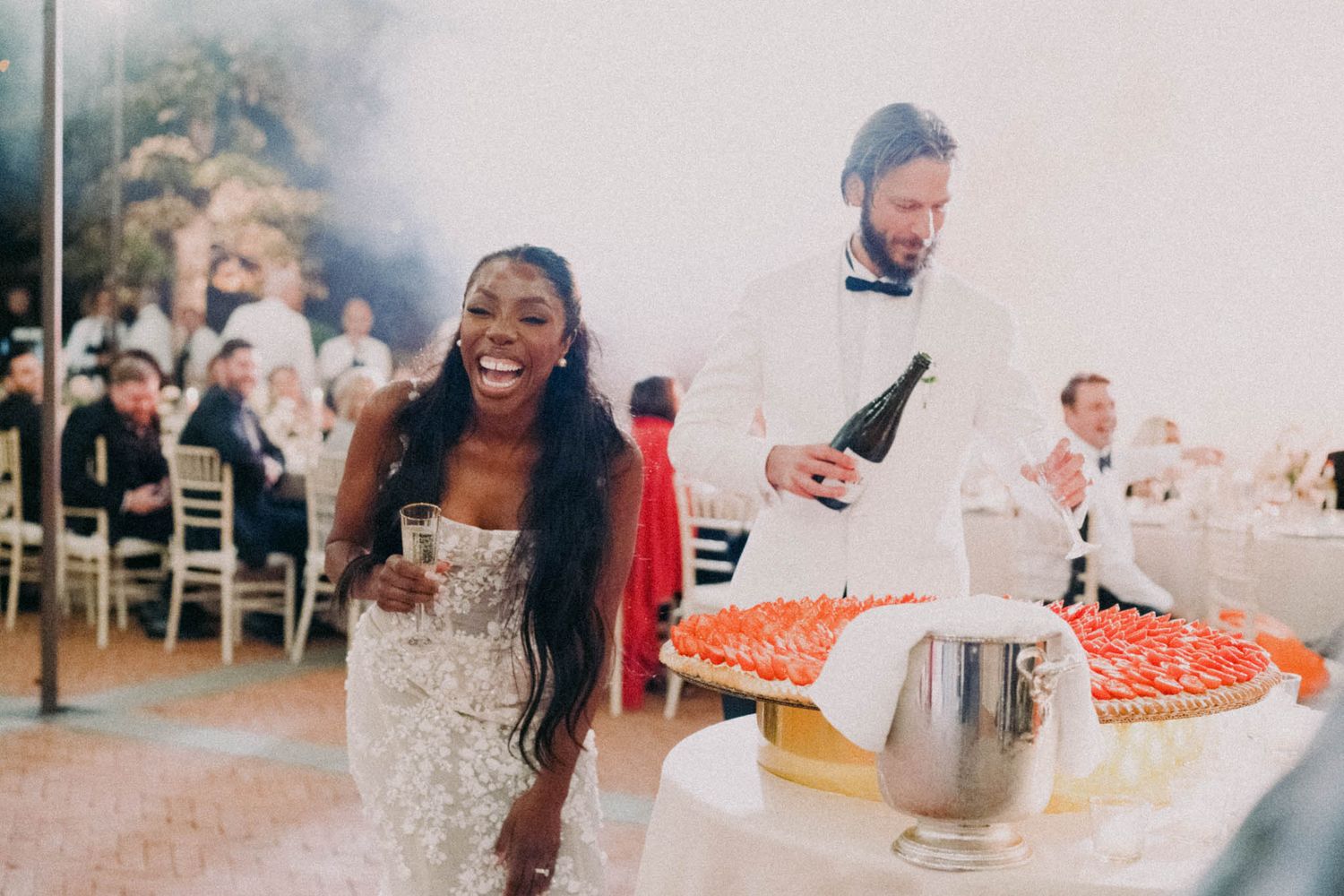 A group celebrates at a party with champagne bottles and pizza slices in a bright indoor setting.