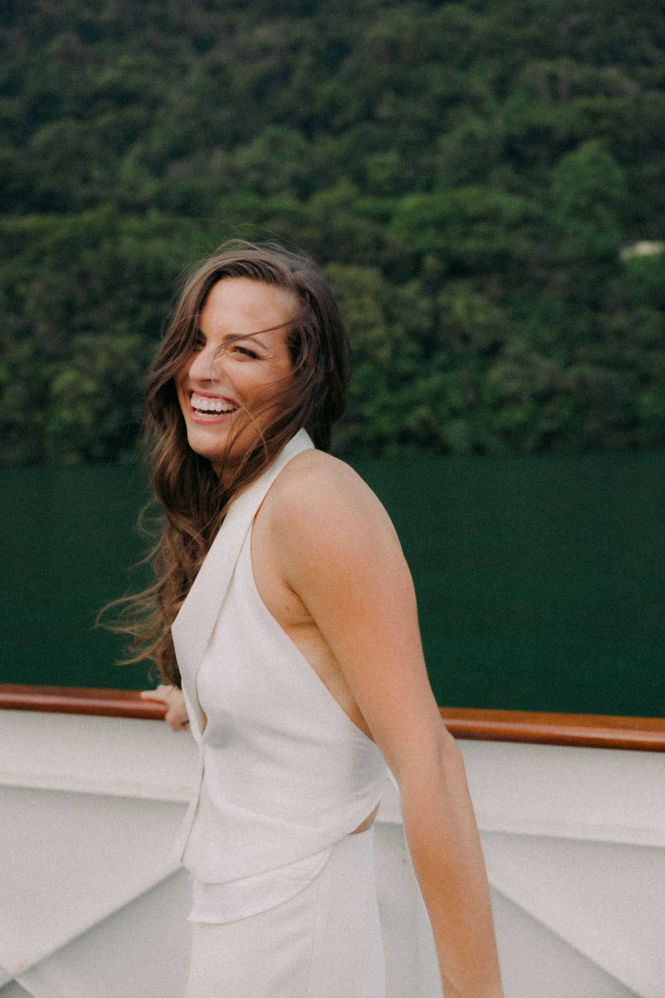 A joyful person in a white sleeveless dress laughs while enjoying time on a boat with green hills in the background.